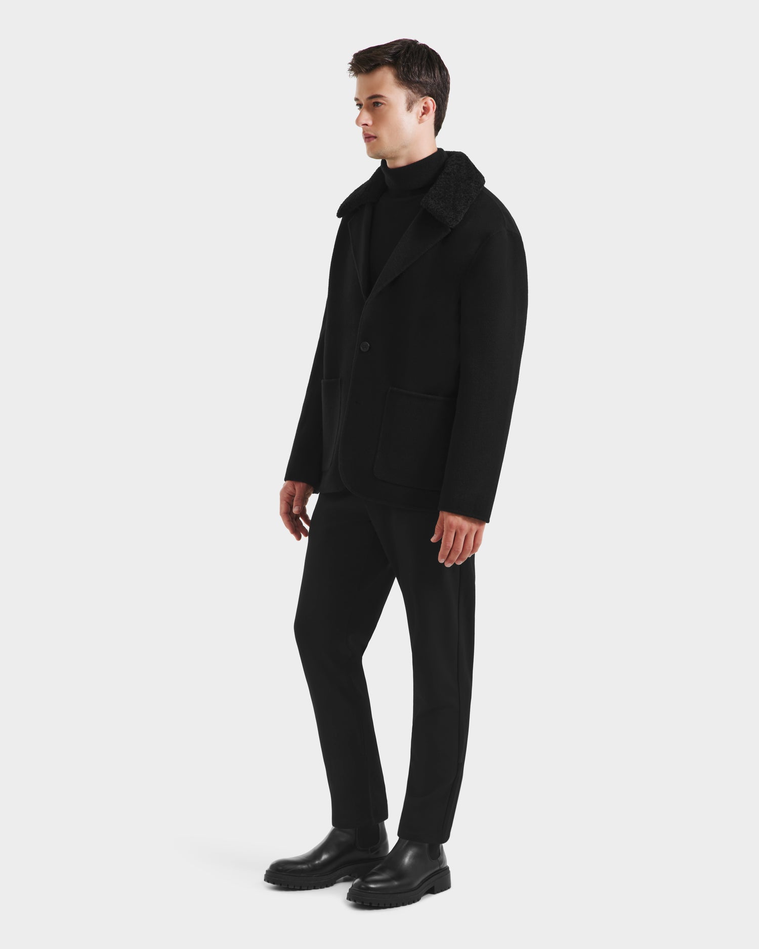 A man stands against a white background wearing the Rudsak AIDEN - 6125558 Black wool-blend coat with removable faux shearling collar, styled with black turtleneck, pants, and shoes. He faces left, hands by his sides.