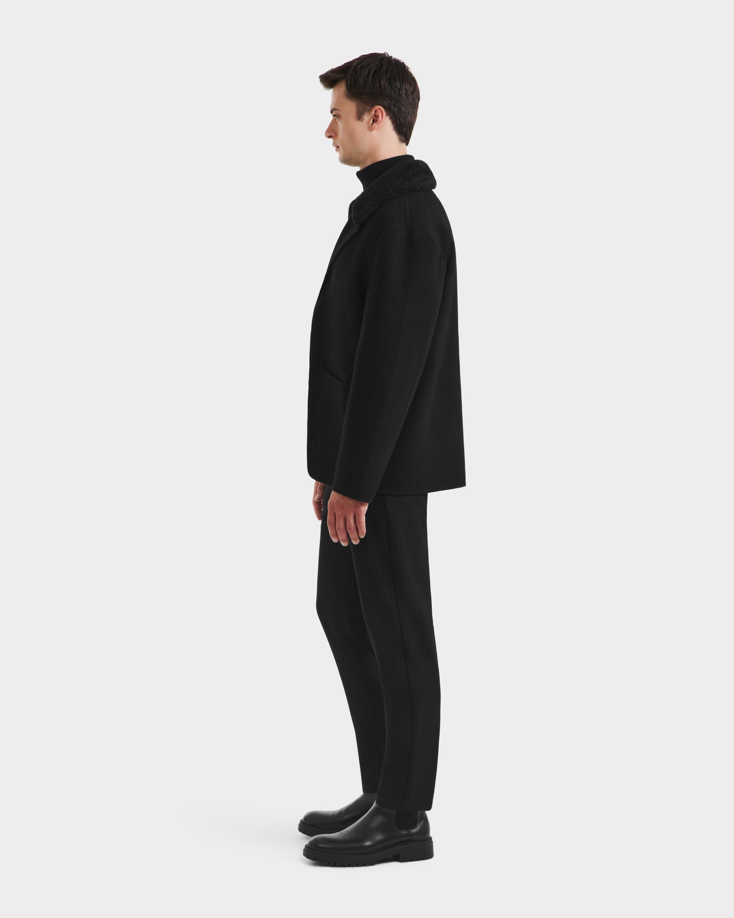 A man stands in profile against a plain white background, wearing the Rudsak AIDEN - 6125558 Black wool-blend coat with a removable faux shearling collar, paired with black pants and shoes, highlighting this refined outerwear piece.