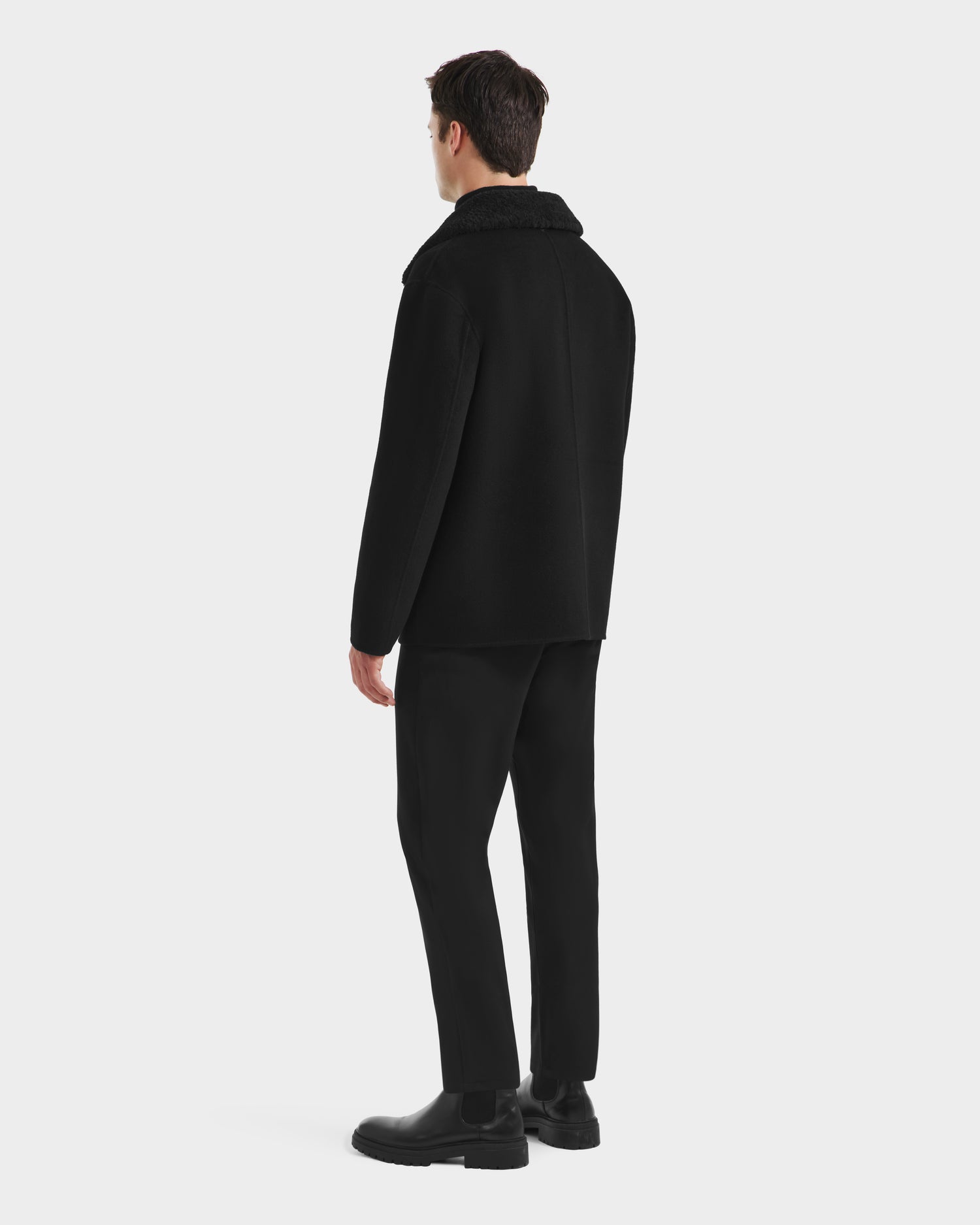 A man faces away, wearing the Rudsak AIDEN - 6125558 Black men’s wool-blend coat with a removable faux shearling collar, paired with black pants and boots, set against a plain white background.