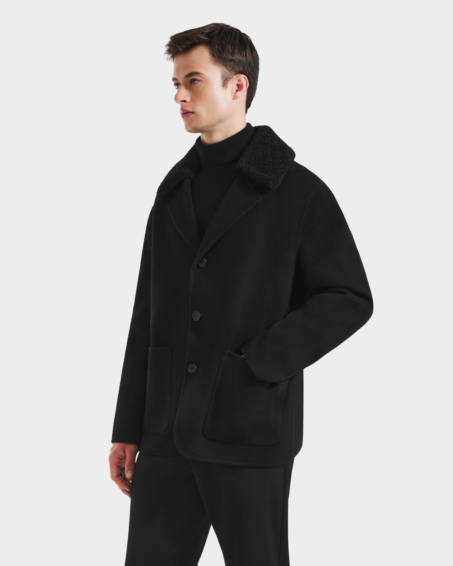 A man wearing the Rudsak AIDEN - 6125558 Black wool-blend coat with large front pockets and a black turtleneck stands against a plain white background, looking to the side.