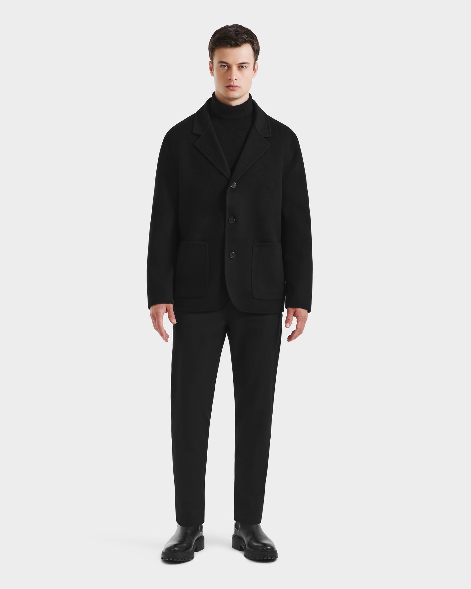 A man faces forward against a plain white background, wearing the Rudsak AIDEN - 6125558 Black men’s wool-blend coat over a black turtleneck sweater, paired with black pants and shoes.