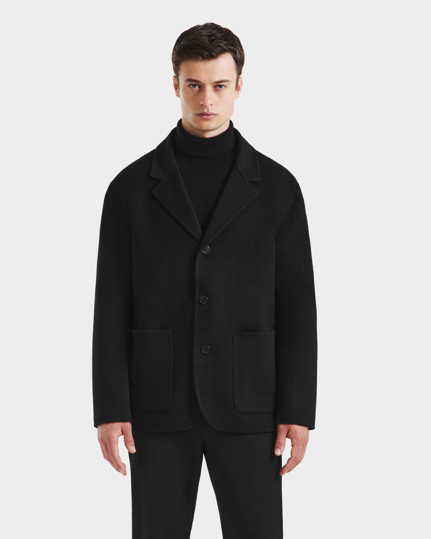 A man stands facing forward with a neutral expression, wearing the Rudsak AIDEN - 6125558 Black men’s wool-blend coat featuring a removable faux shearling collar over a black turtleneck and pants, set against a plain white background.