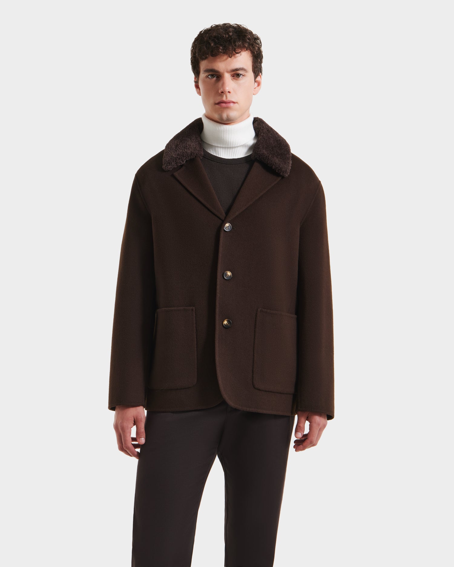 A man stands facing forward against a plain white background, wearing Rudsaks AIDEN - 6125558 Espresso mens wool-blend coat with removable faux shearling collar, paired with a white turtleneck sweater and dark pants.