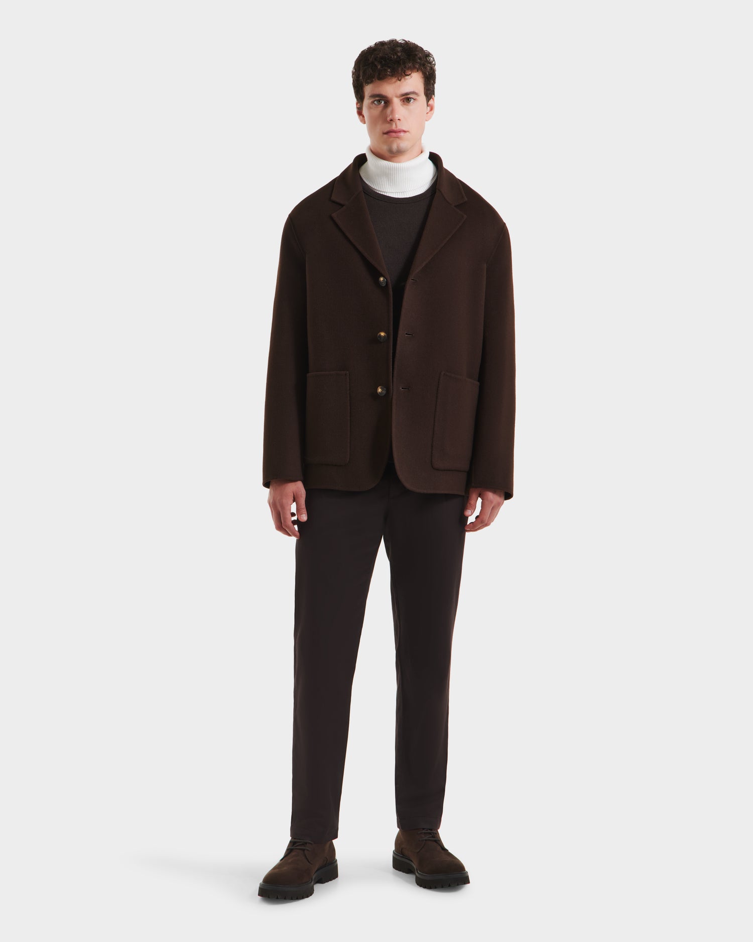 A man stands facing forward in a refined look, wearing the Rudsak AIDEN - 6125558 Espresso men’s wool-blend coat with two front pockets, paired with dark brown pants, a white turtleneck sweater, and dark shoes against a plain white background.