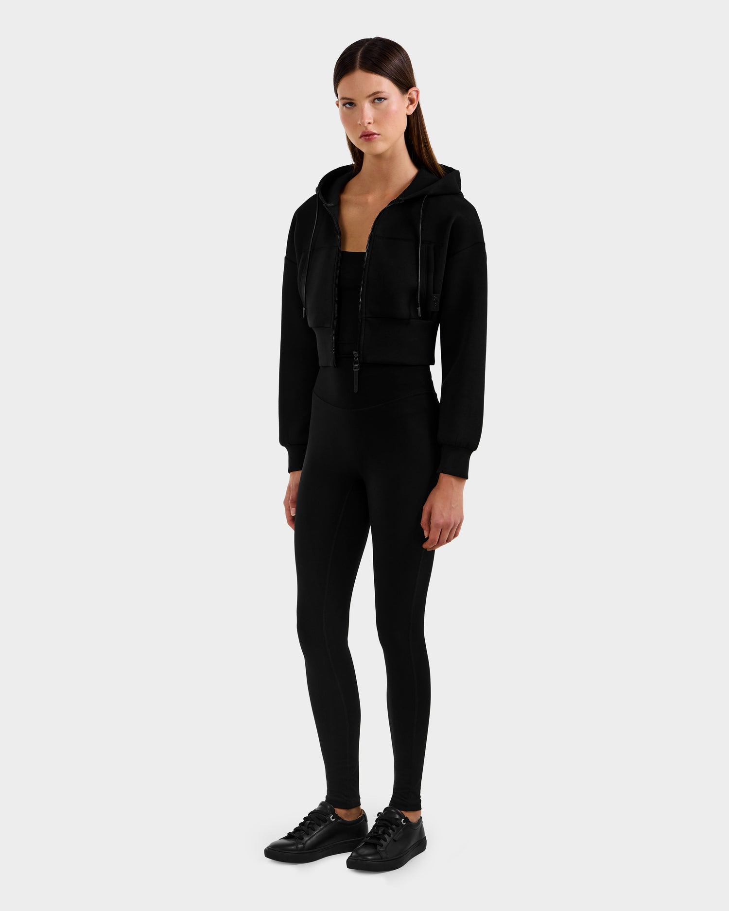 A woman faces forward wearing the Rudsak ALAYA - 8525038 Black cropped hoodie, black leggings, and sneakers against a light gray background. Her straight long brown hair and neutral expression complete the casual-chic look.