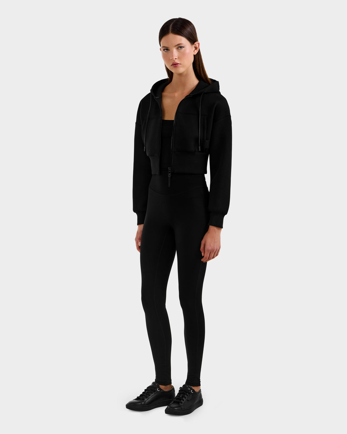 A woman faces forward wearing the Rudsak ALAYA - 8525038 Black cropped hoodie, black leggings, and sneakers against a light gray background. Her straight long brown hair and neutral expression complete the casual-chic look.