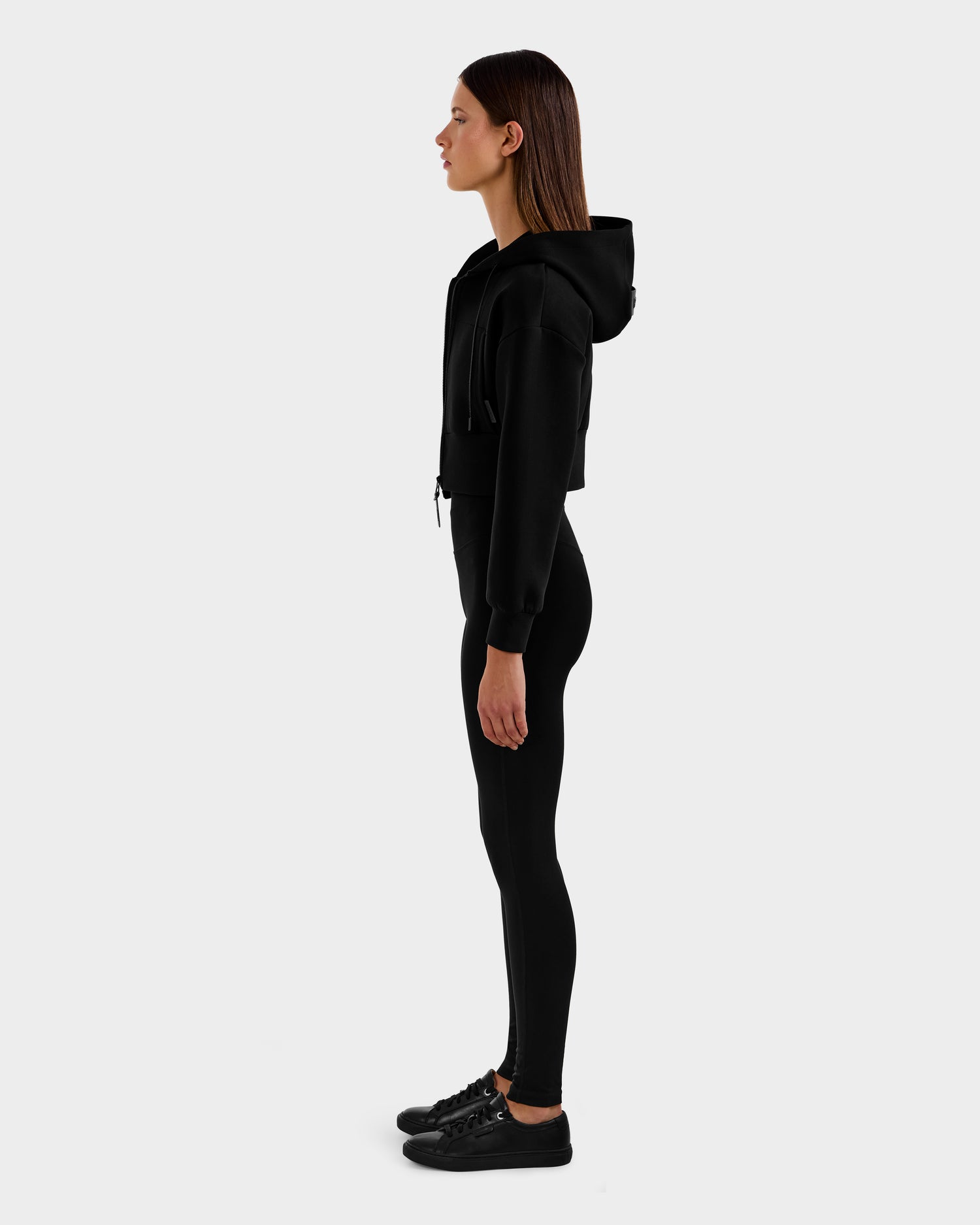 A woman in profile faces left, wearing the Rudsak ALAYA - 8525038 Black cropped hoodie with black leggings and sneakers, posing against a plain light gray background for a sleek, casual-chic loungewear look.
