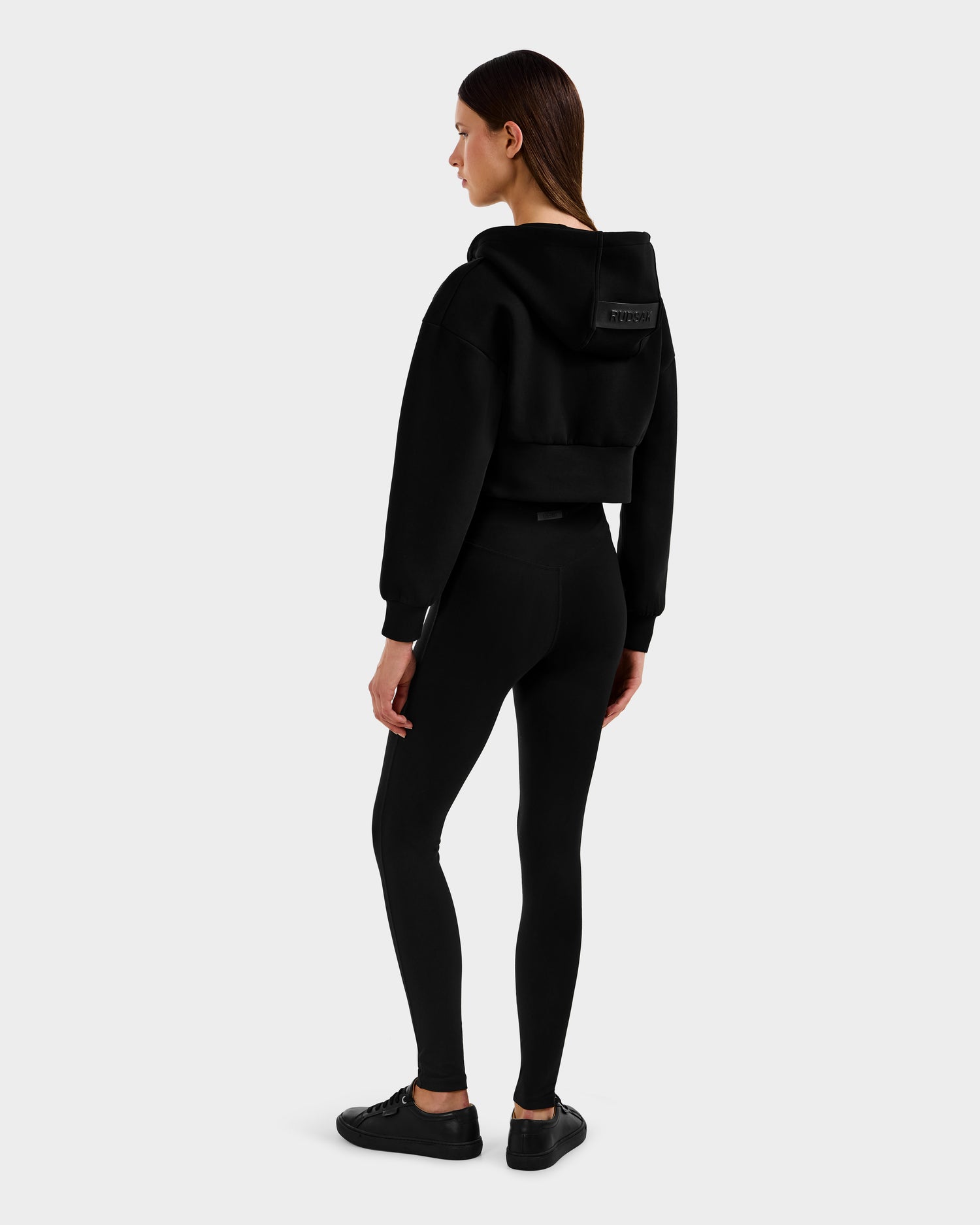 A woman stands sideways, wearing the Rudsak ALAYA - 8525038 Black cropped hoodie with matching leggings and sneakers, capturing a casual-chic loungewear vibe against a plain light gray background.