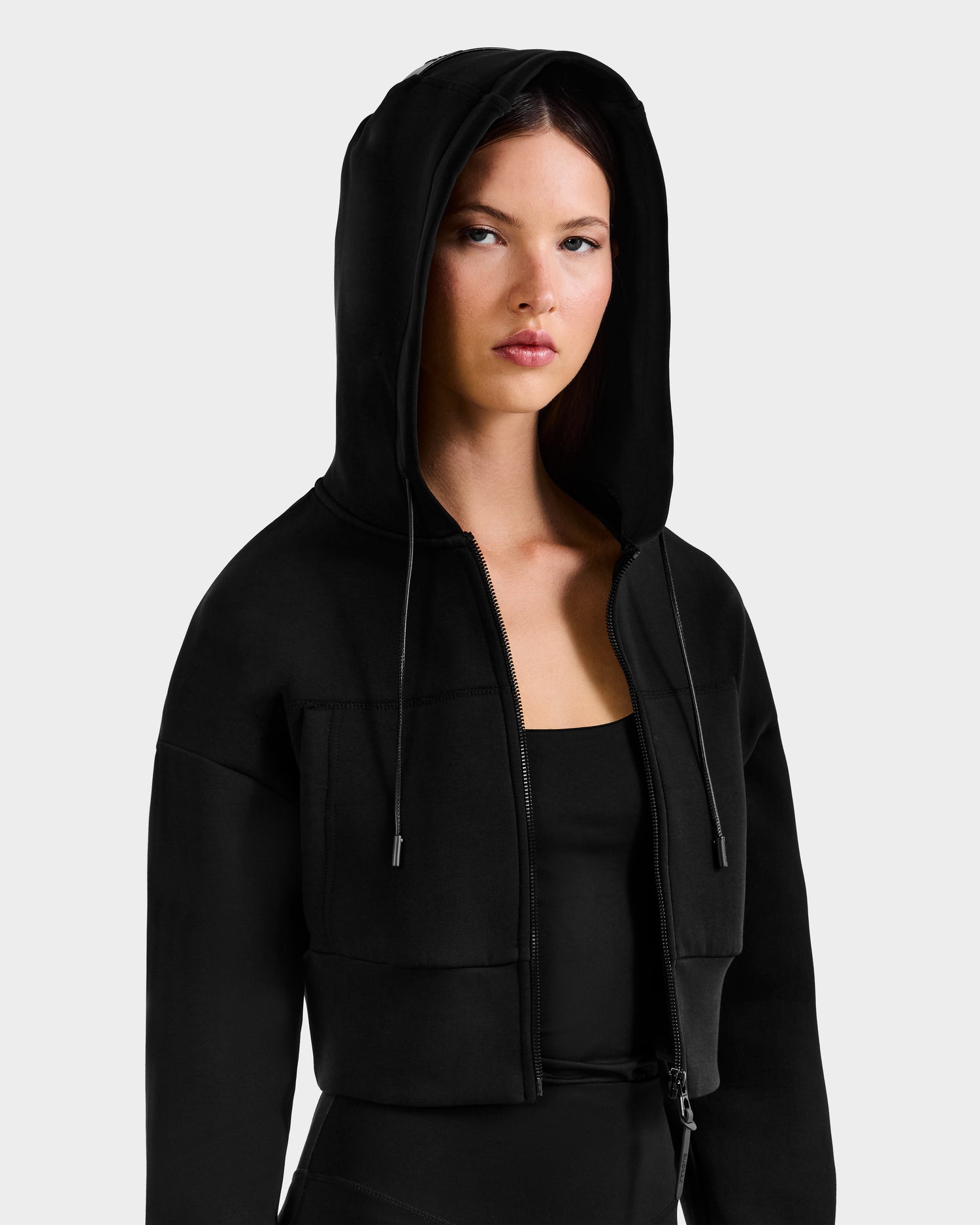 A woman with straight brown hair wears the Rudsak ALAYA - 8525038 Black zip-up hoodie over a black top, hood up, looking forward neutrally and embodying a casual-chic loungewear vibe against a plain light background.
