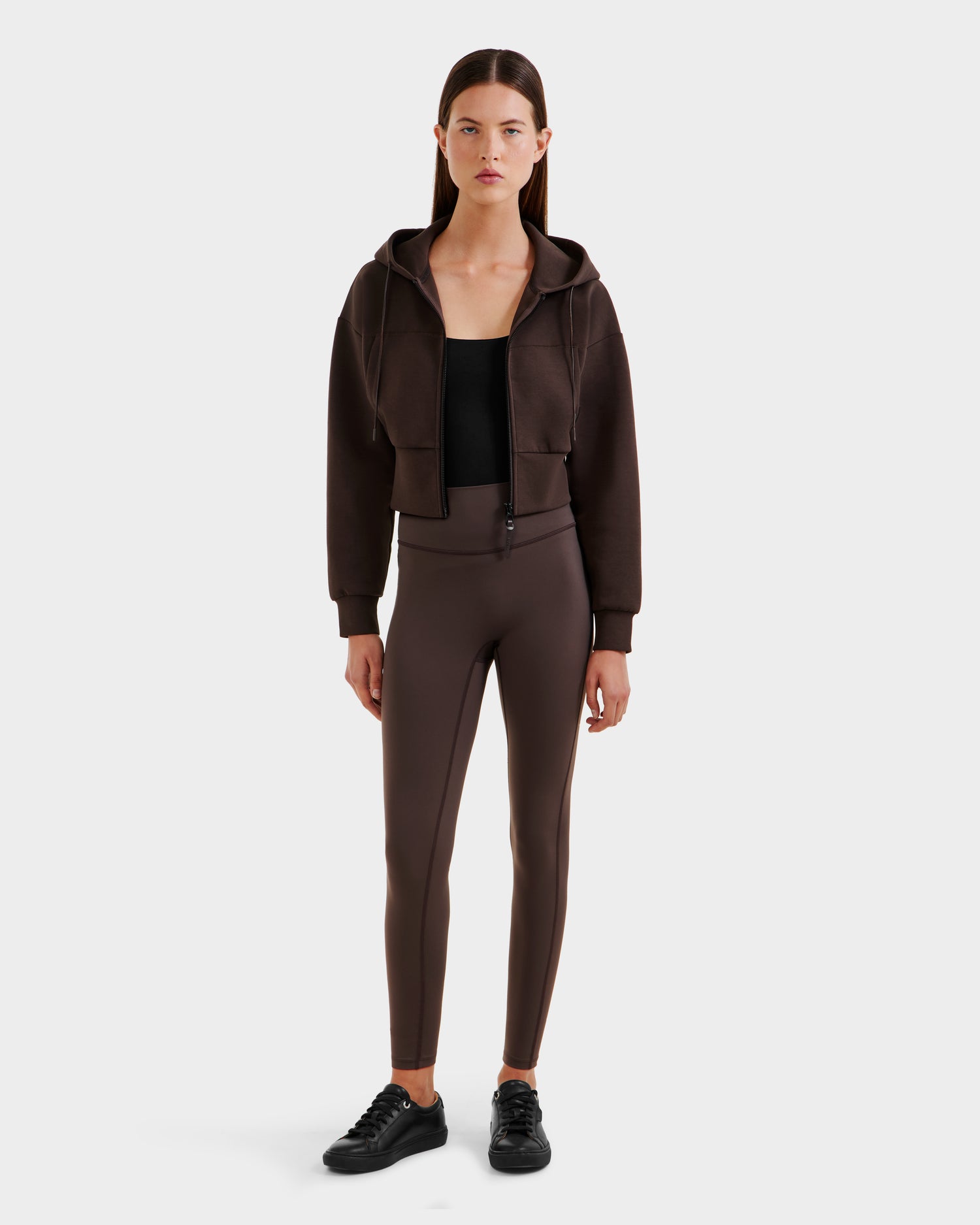 A woman with straight brown hair and a neutral expression wears the ALAYA - 8525038 Espresso cropped zip-up hoodie by Rudsak over a black top, paired with brown high-waisted leggings and black sneakers against a plain background.
