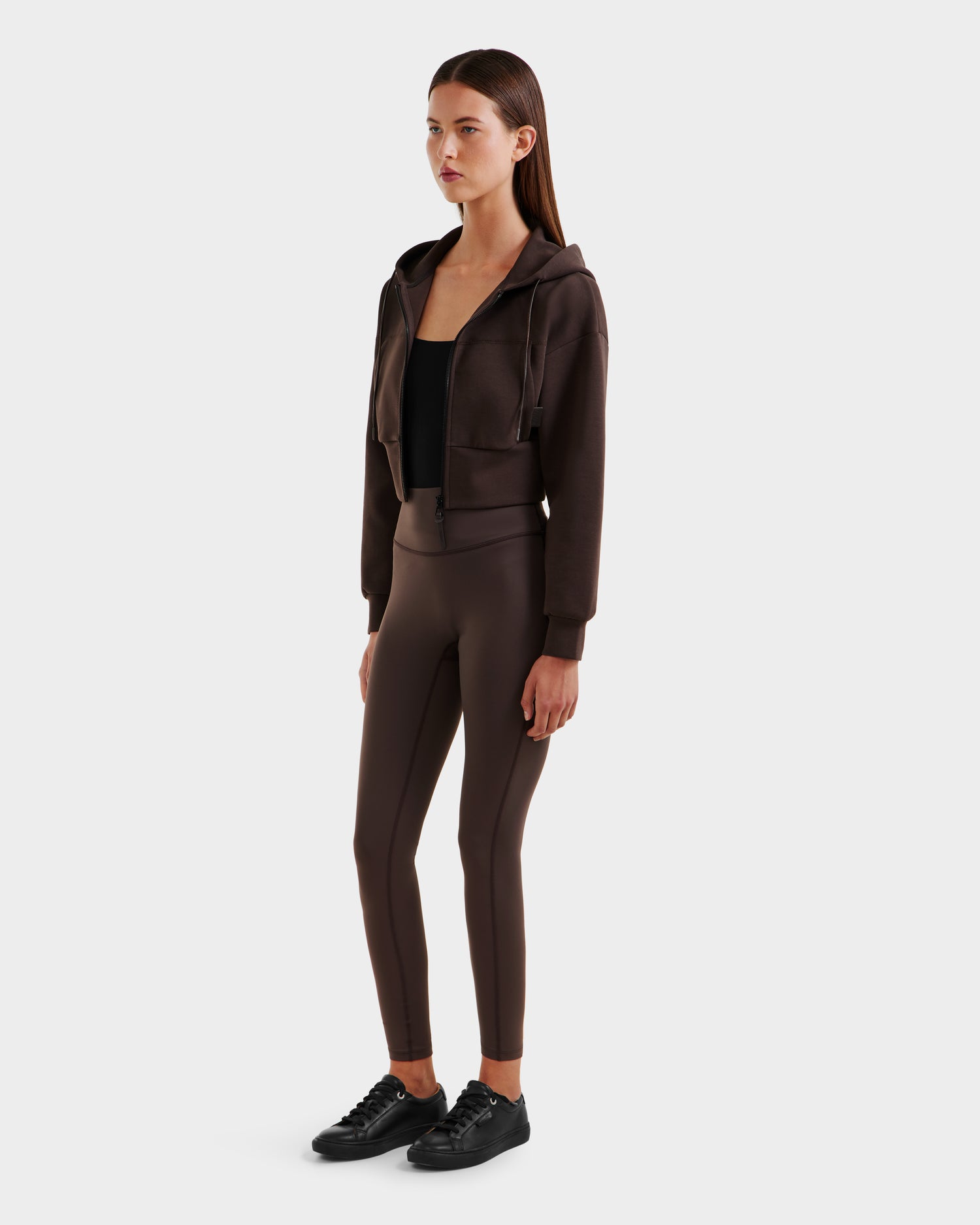 A woman faces slightly to the side, wearing the Rudsak ALAYA - 8525038 Espresso double jersey cropped zip-up hoodie with matching brown high-waisted leggings, black tank top, and black sneakers against a plain light background.