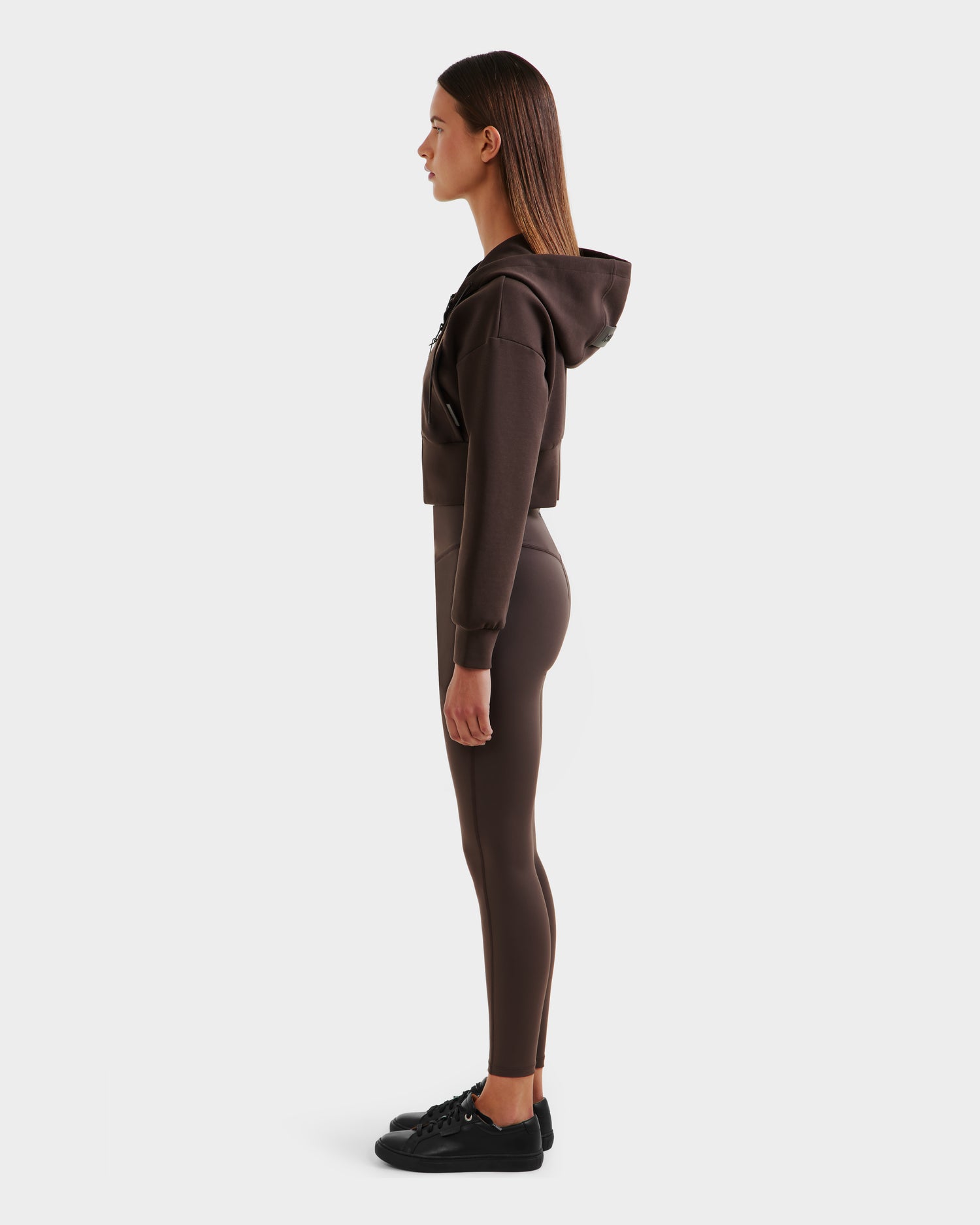 A woman stands in profile against a light gray background, wearing the Rudsak ALAYA - 8525038 Espresso women’s cropped zip-up hoodie and matching leggings in premium double jersey, paired with black sneakers.
