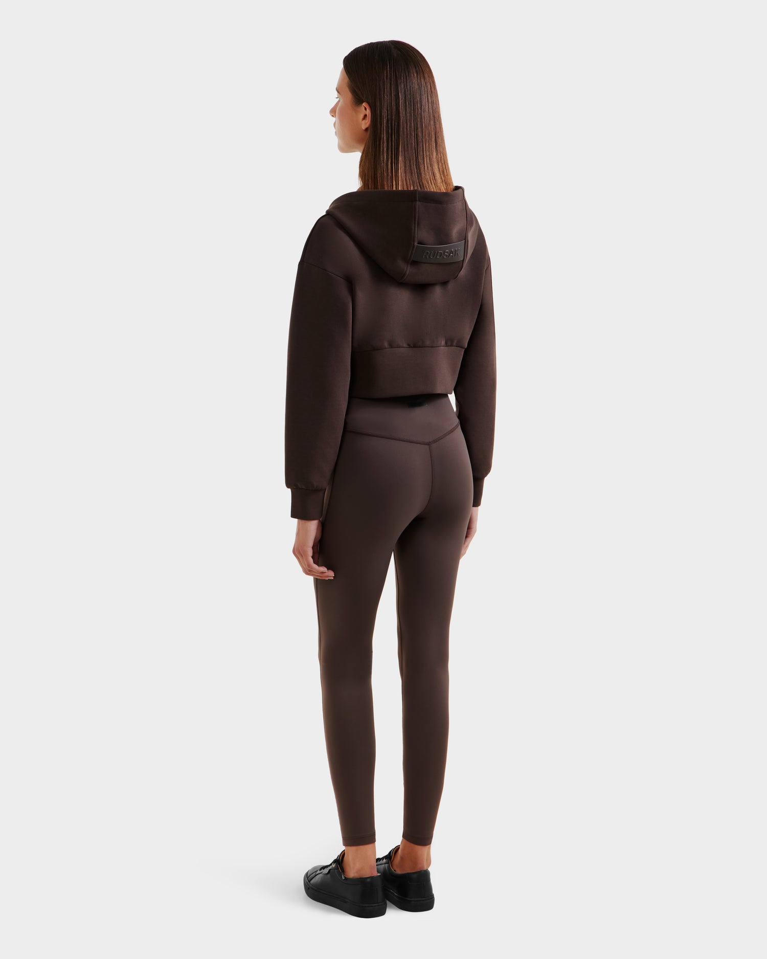A woman faces away, styled in the Rudsak ALAYA - 8525038 Espresso cropped zip-up hoodie and matching fitted leggings, paired with black sneakers, against a plain light gray background.