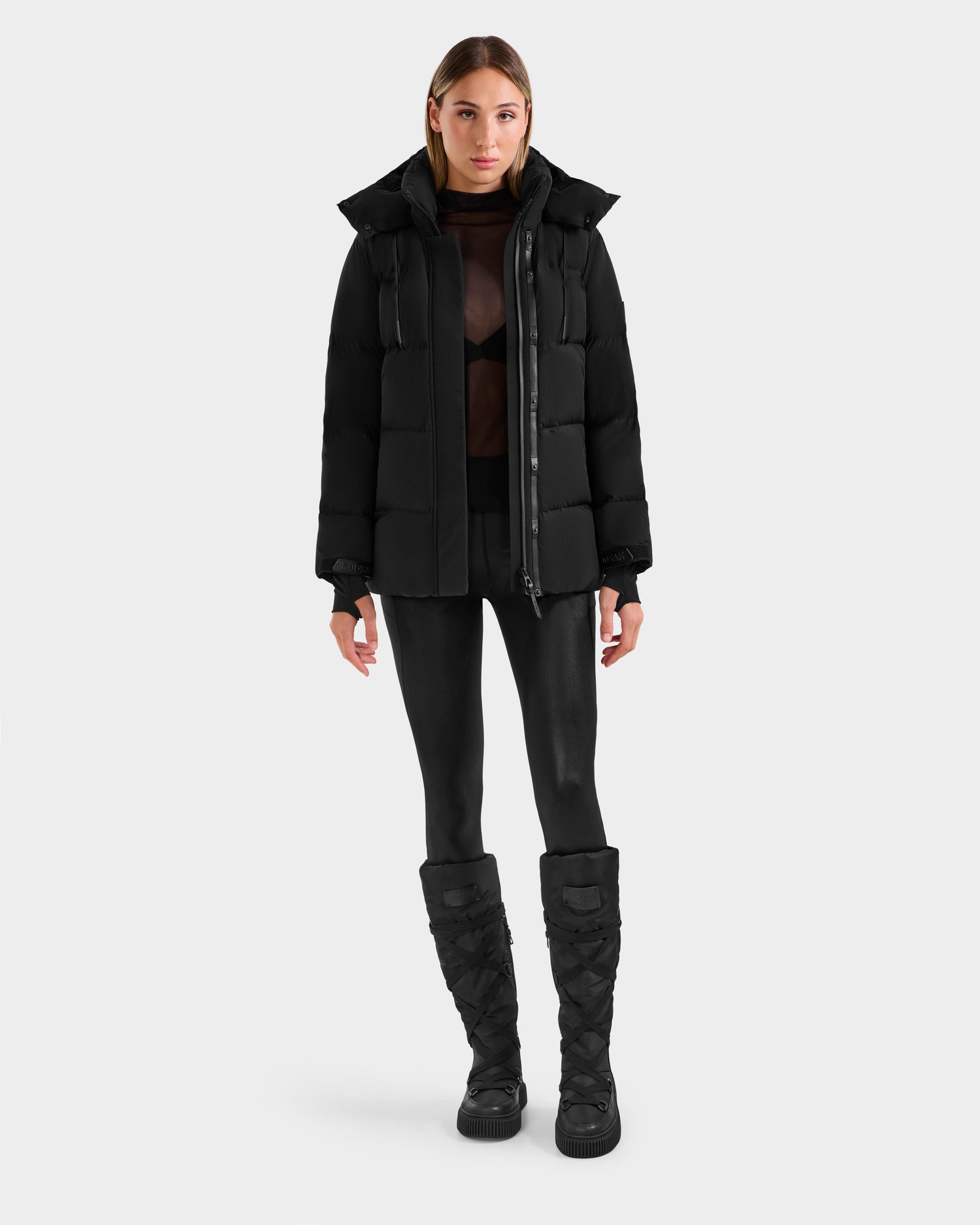 A woman faces forward in a Rudsak ALESSANDRA S-M - L125551 Black cropped down puffer jacket with removable hood, styled with a sheer black top, black leggings, and quilted boots against a light gray background.