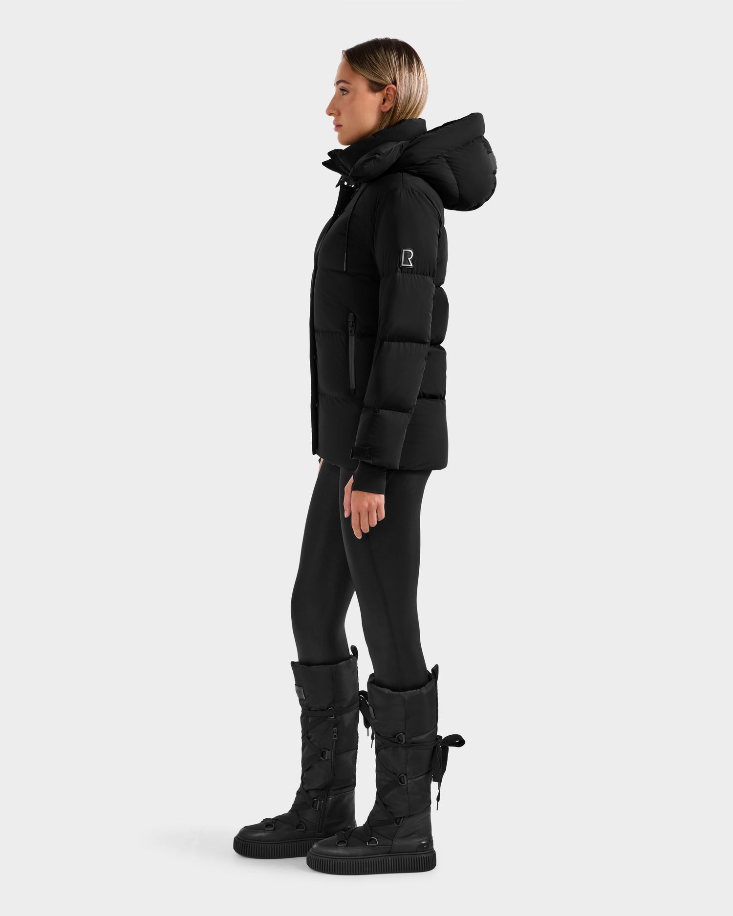 A person stands in profile wearing the Rudsak ALESSANDRA S-M - L125551 Black cropped down puffer with removable hood, styled with black leggings and winter boots against a light gray background—a cold-weather essential.