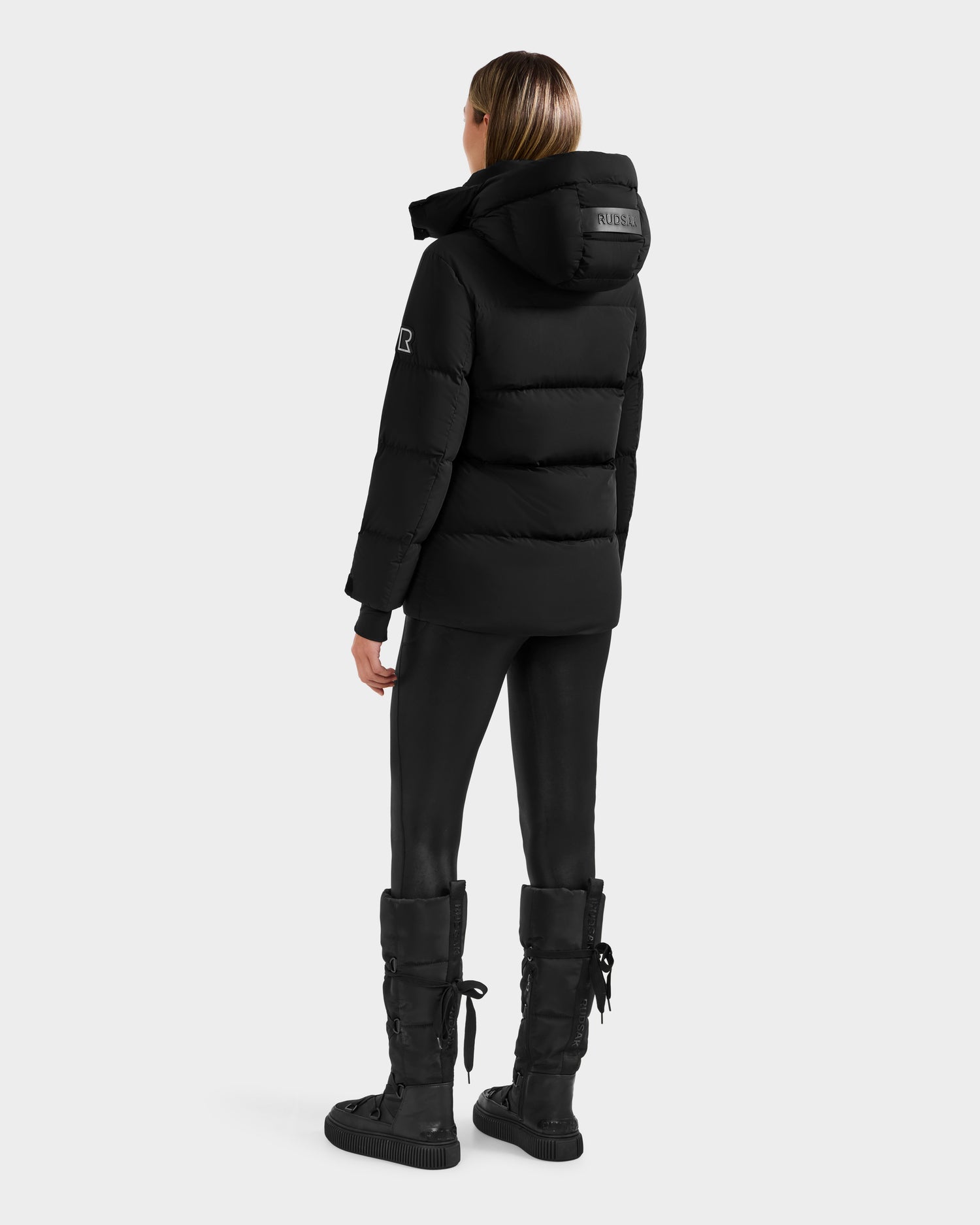 A person stands facing away, wearing the Rudsak ALESSANDRA S-M - L125551 Black cropped down puffer jacket with logo patches on the sleeve and hood, styled with black leggings and lace-up boots against a plain white background.