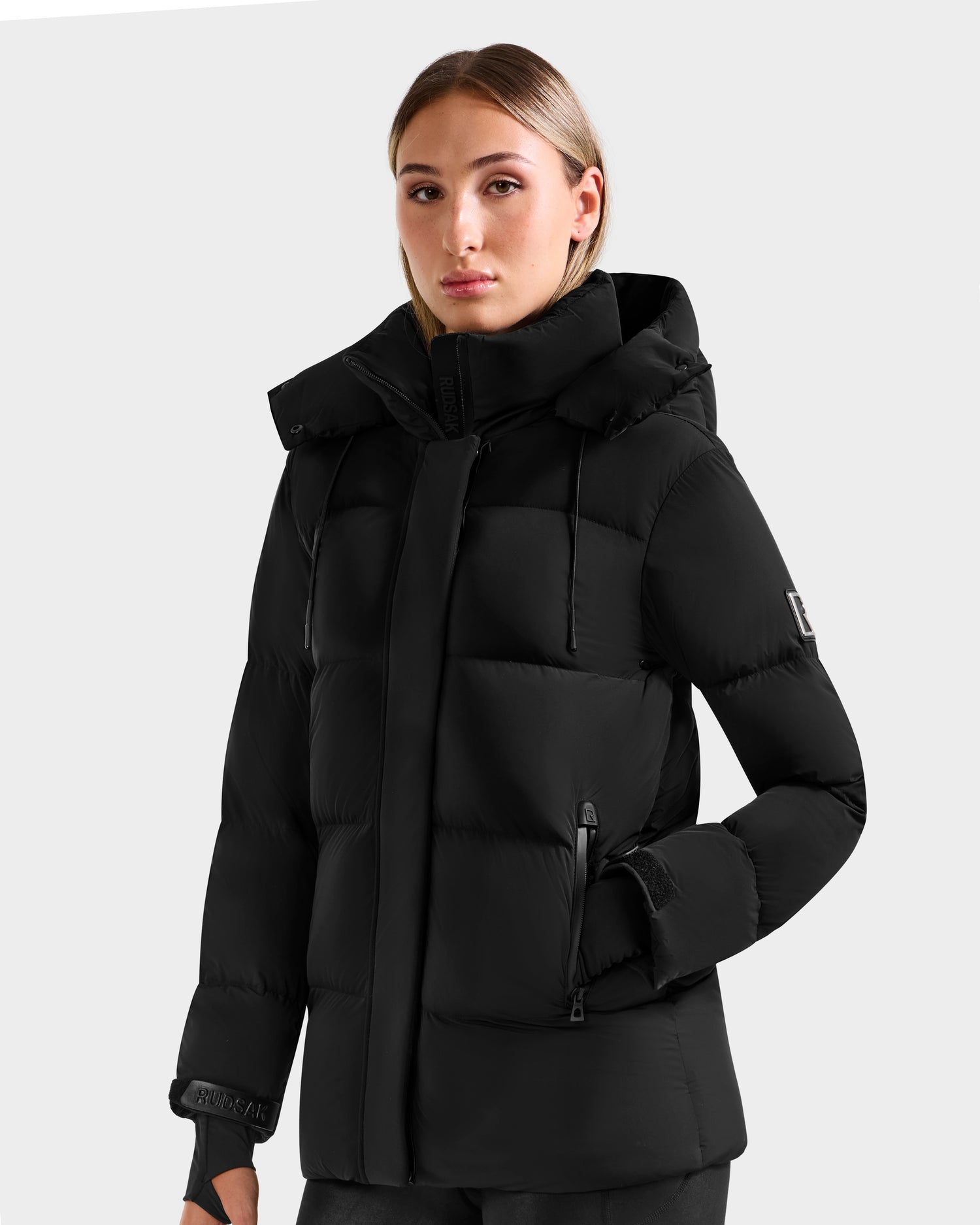 A person with long straight hair wears the Rudsak ALESSANDRA S-M - L125551 Black down puffer jacket, featuring a hood, zippered front, and side pockets, standing against a plain light gray background.