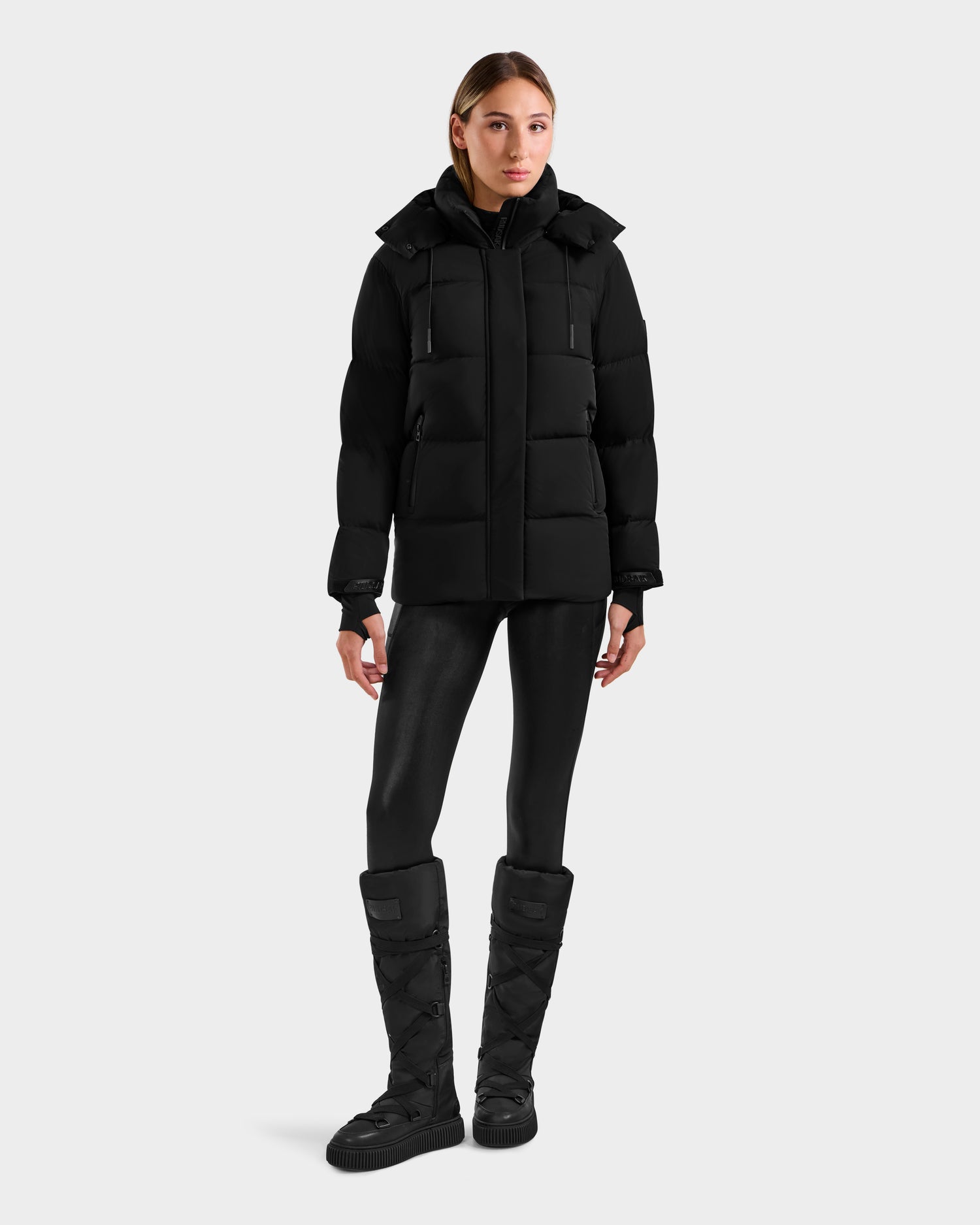 A woman faces forward in a Rudsak ALESSANDRA S-M - L125551 Black puffer jacket with removable hood, paired with black shiny leggings and black padded knee-high boots against a plain light gray background.