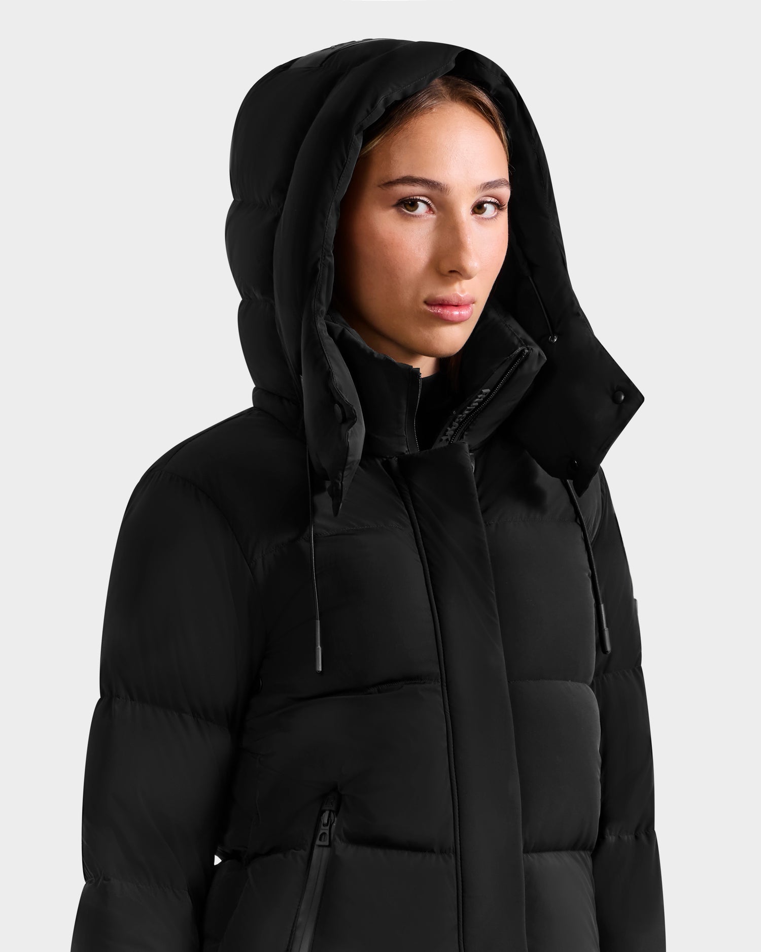 A young woman wears the Rudsak ALESSANDRA S-M - L125551 Black cropped down puffer with a removable hood up, looking slightly to the side against a plain light grey background—her cold-weather essential.