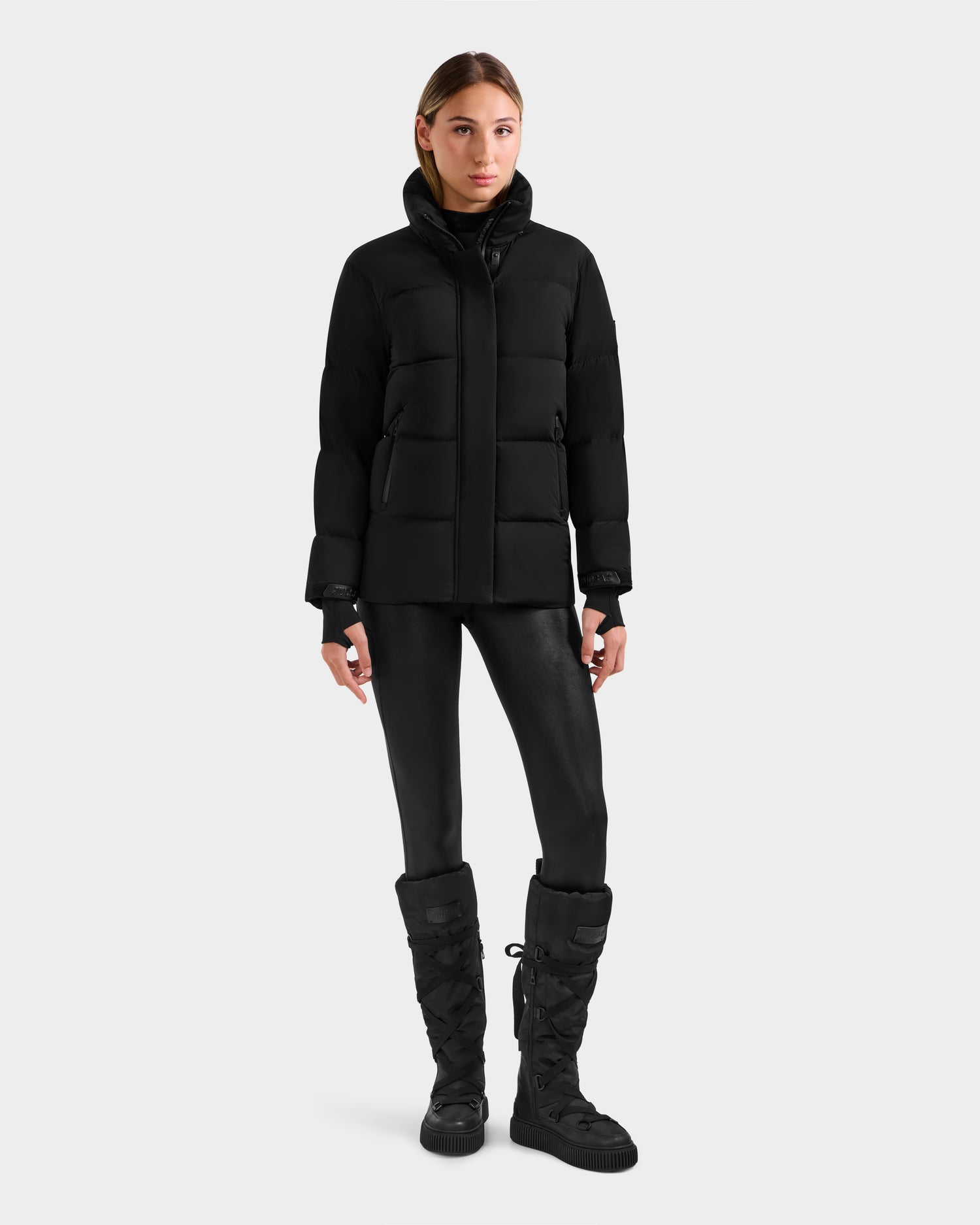 A woman stands facing forward against a light gray background, wearing the Rudsak ALESSANDRA S-M - L125551 Black cropped down puffer jacket with removable hood—an effortlessly chic cold-weather essential.