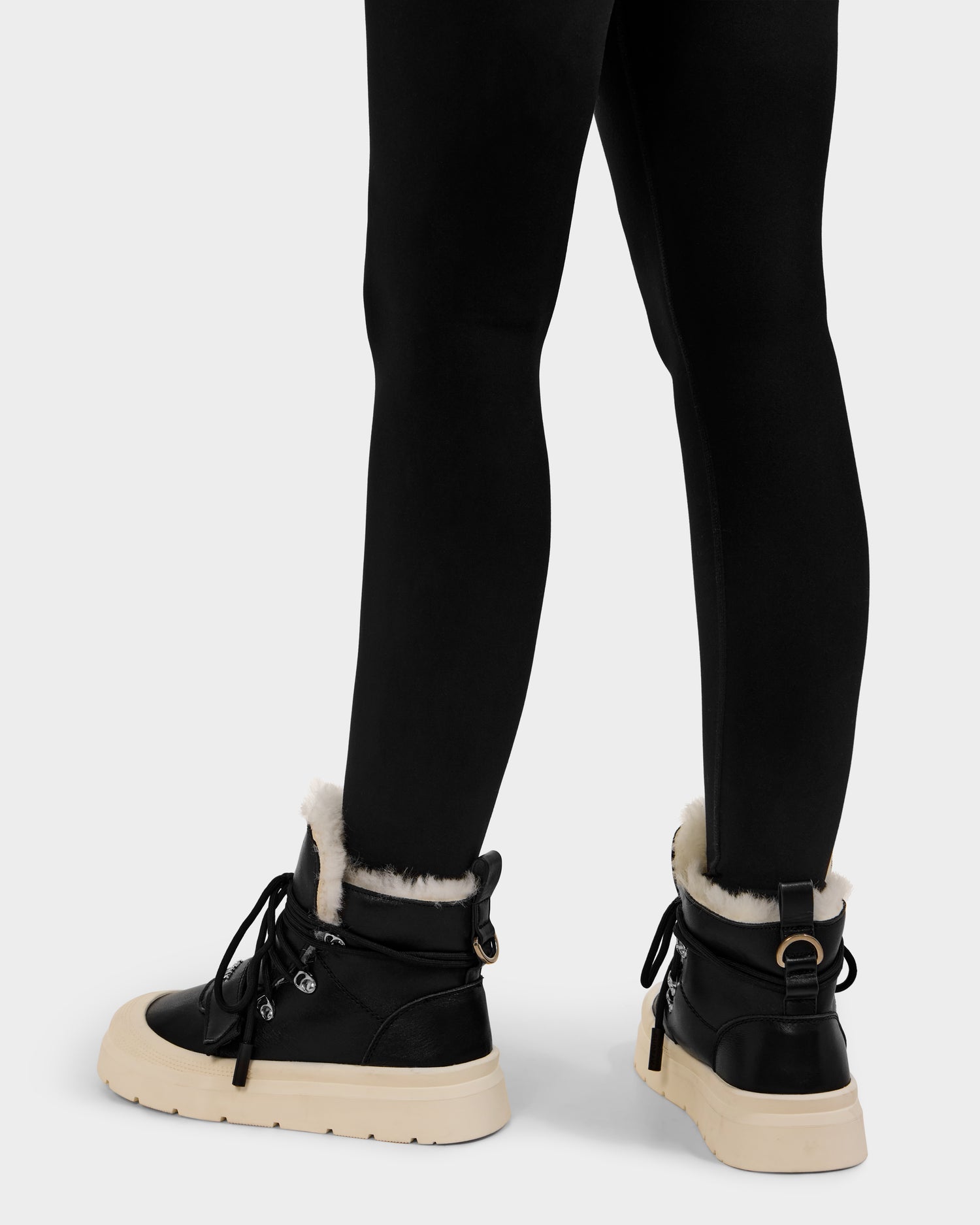A person shown from the back wears Rudsak ALEXEY L - L225518 Black women’s leather boots with white soles and soft wool lining, paired with black leggings, against a plain light background.