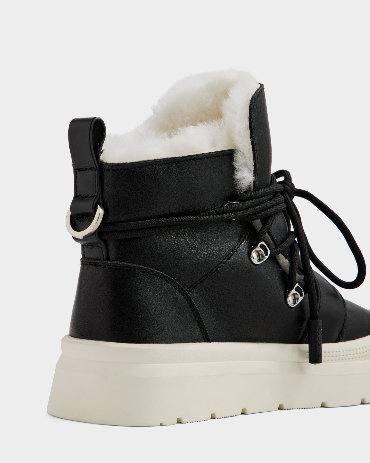 A close-up of Rudsak’s ALEXEY L - L225518 Black women’s leather boot featuring black laces, metal eyelets, plush wool lining, and a thick white rubber sole set against a neutral background.