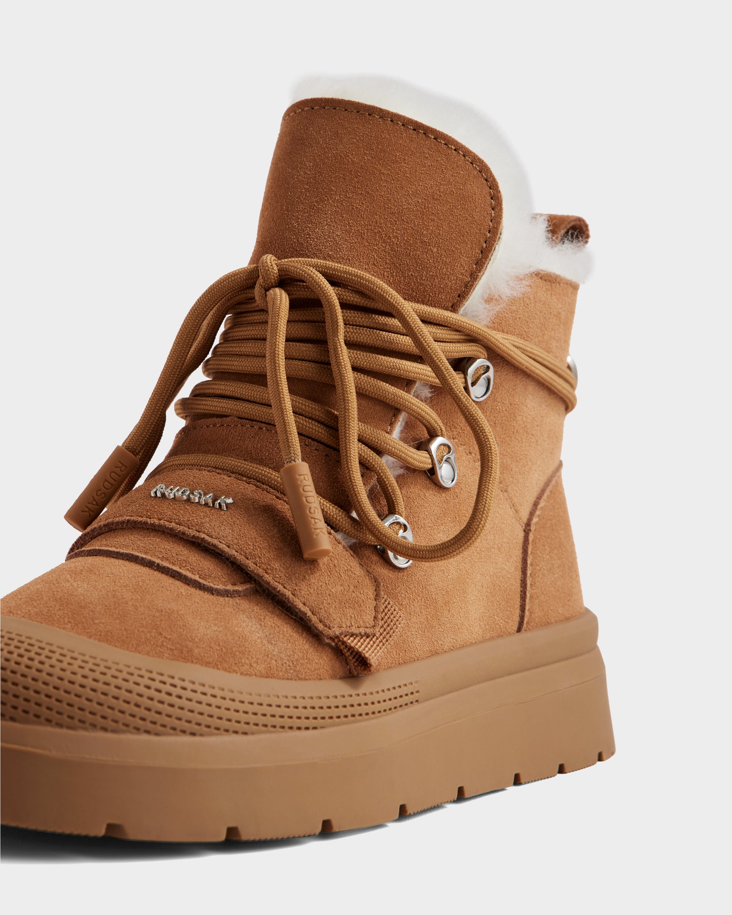 A close-up of Rudsak’s ALEXEY - L225025 Tan women's suede winter boot with thick laces, metal eyelets, a gum-colored traction sole, and plush white wool lining visible at the top.