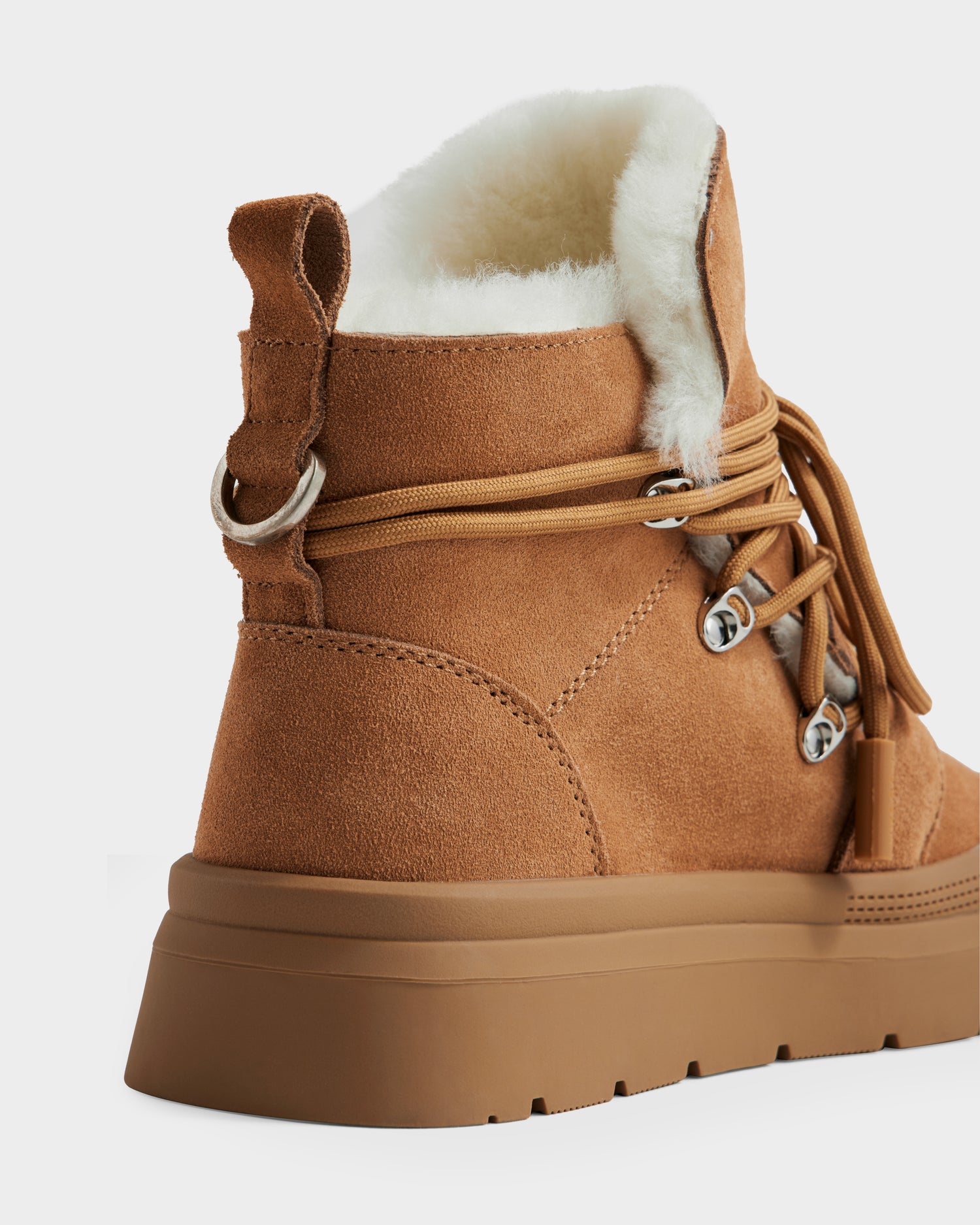 A close-up of the Rudsak ALEXEY - L225025 Tan women’s suede winter boot, featuring a thick rubber sole for durable traction, metal lace hooks, beige laces, and soft white fleece lining at the ankle.