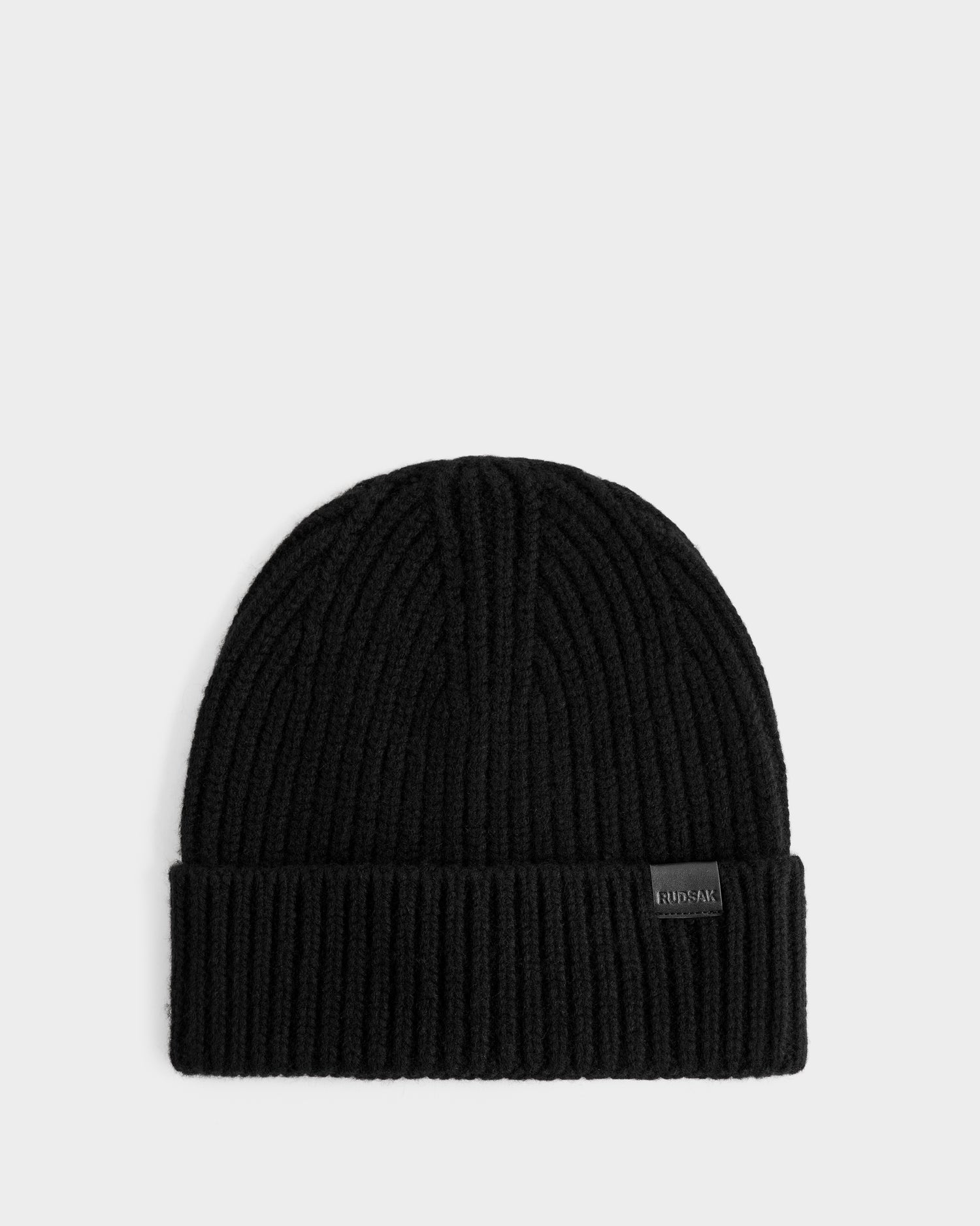 The Rudsak ALINA - L425540 Black is a black ribbed knit winter hat with a folded cuff and a small rectangular front tag, shown against a plain light gray background.