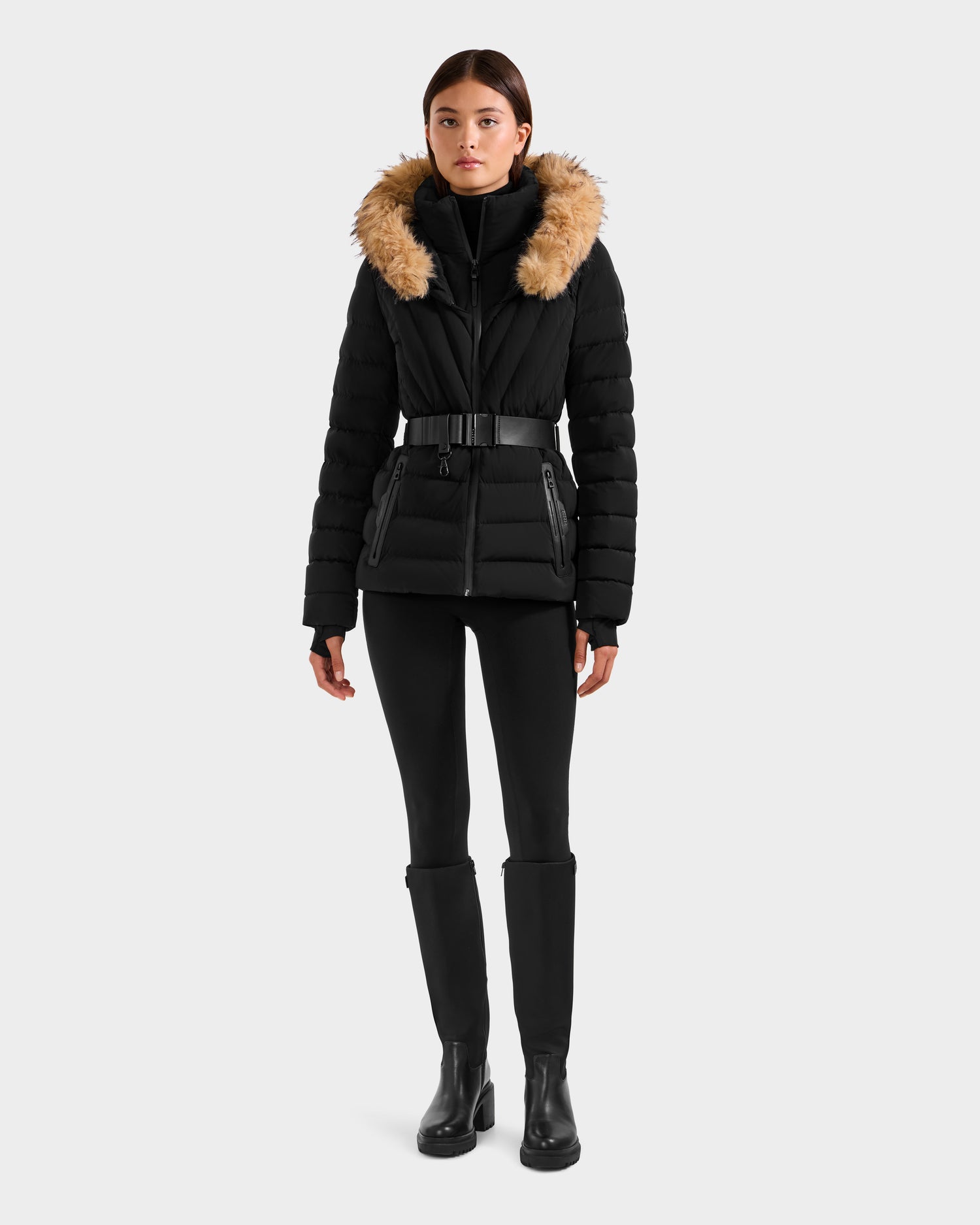 A woman models the Rudsak ALISA F - L125527 Black belted quilted puffer jacket with a brown faux-fur trim hood, styled with black leggings, a turtleneck, and boots against a plain light background.