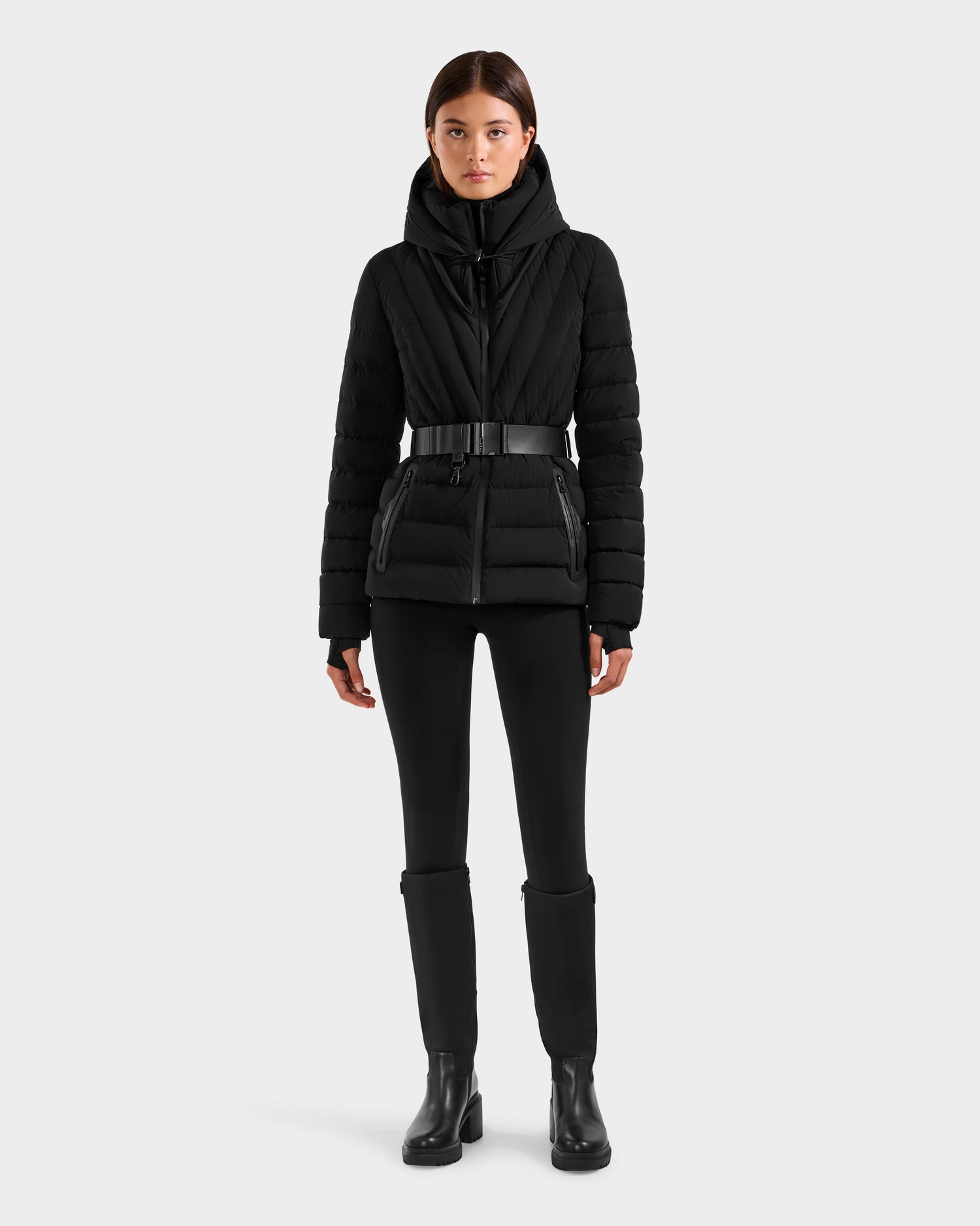 A person stands facing forward wearing the Rudsak ALISA F - L125527 Black quilted puffer jacket with a belt, paired with black pants and ankle boots against a plain light gray background.