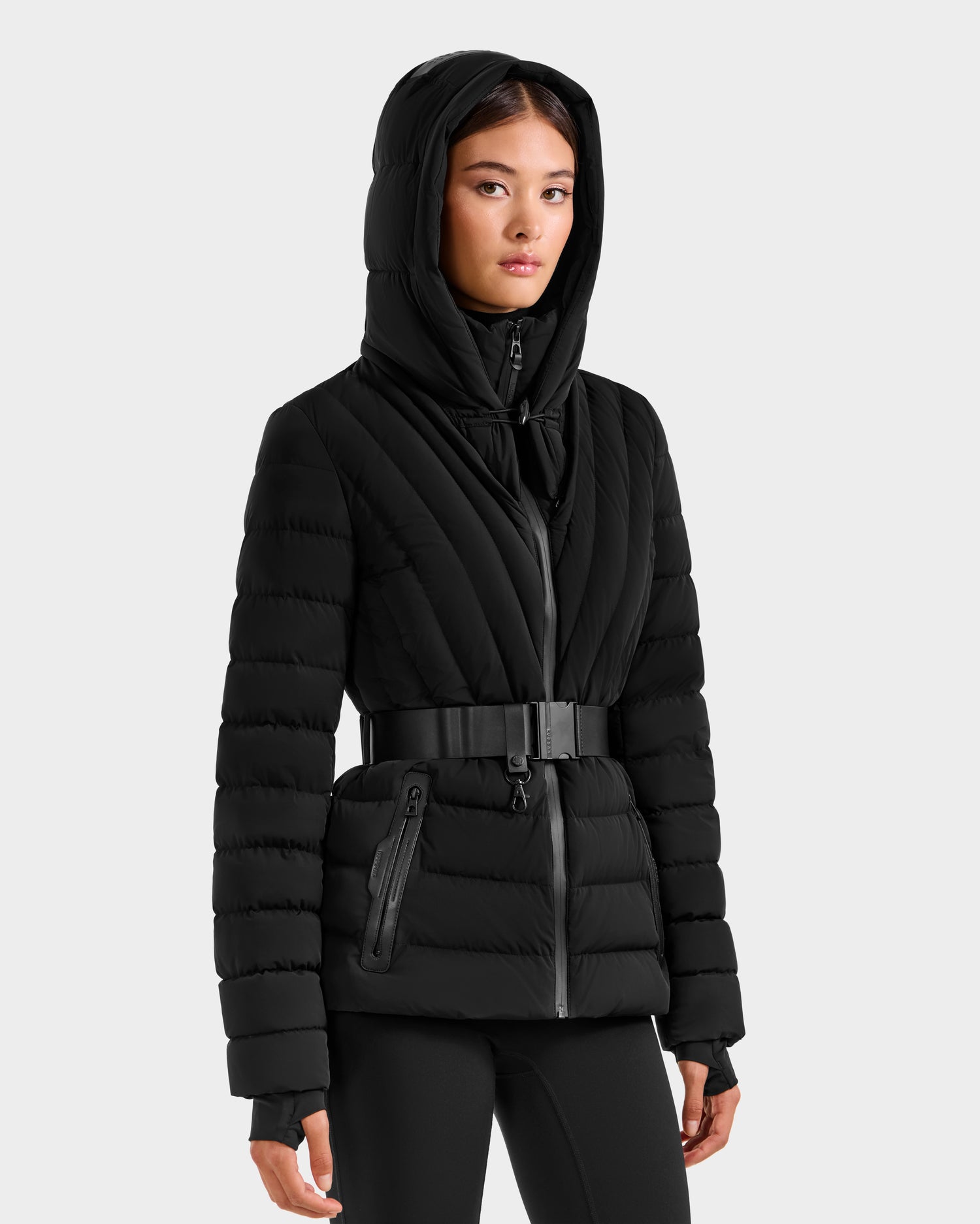 A woman wearing the Rudsak ALISA F - L125527 Black quilted puffer jacket with a belted waist and zipper pockets, paired with black pants, stands against a plain light background.