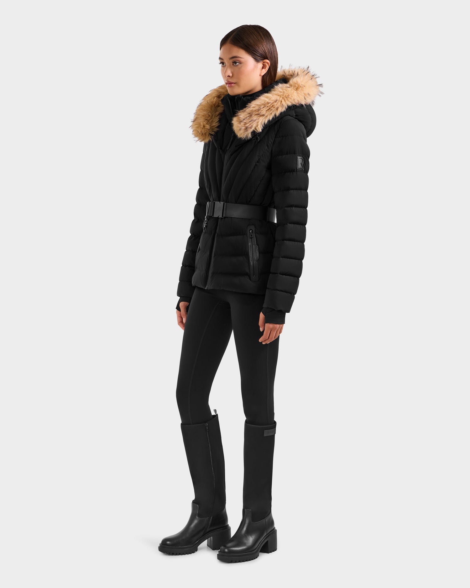 A woman wears the Rudsak ALISA F - L125527 Black quilted puffer jacket with a tan faux-fur trim hood, styled with black leggings and knee-high boots, standing against a plain light gray background.