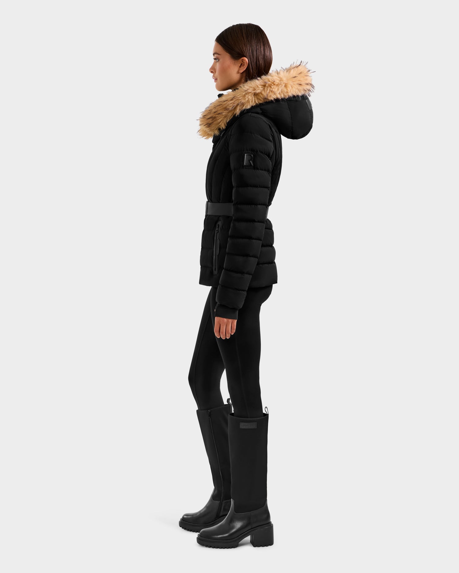 Woman in profile wearing the Rudsak ALISA F - L125527 Black quilted puffer jacket with RDS-certified down and faux-fur trim hood, paired with black leggings and knee-high boots, standing against a plain light gray background.