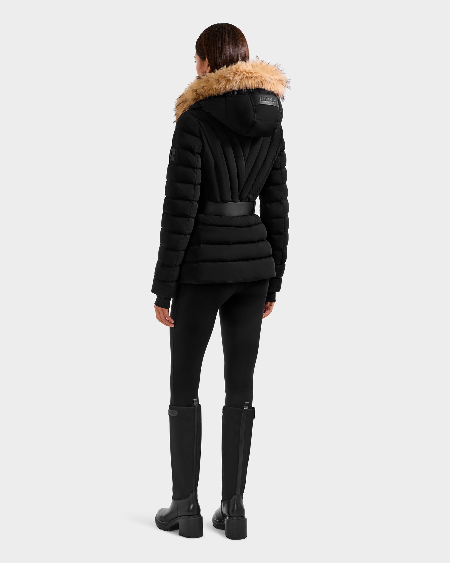 A person faces away wearing the Rudsak ALISA F - L125527 Black quilted puffer jacket with faux-fur trim hood, paired with black pants and tall black boots against a plain light background.