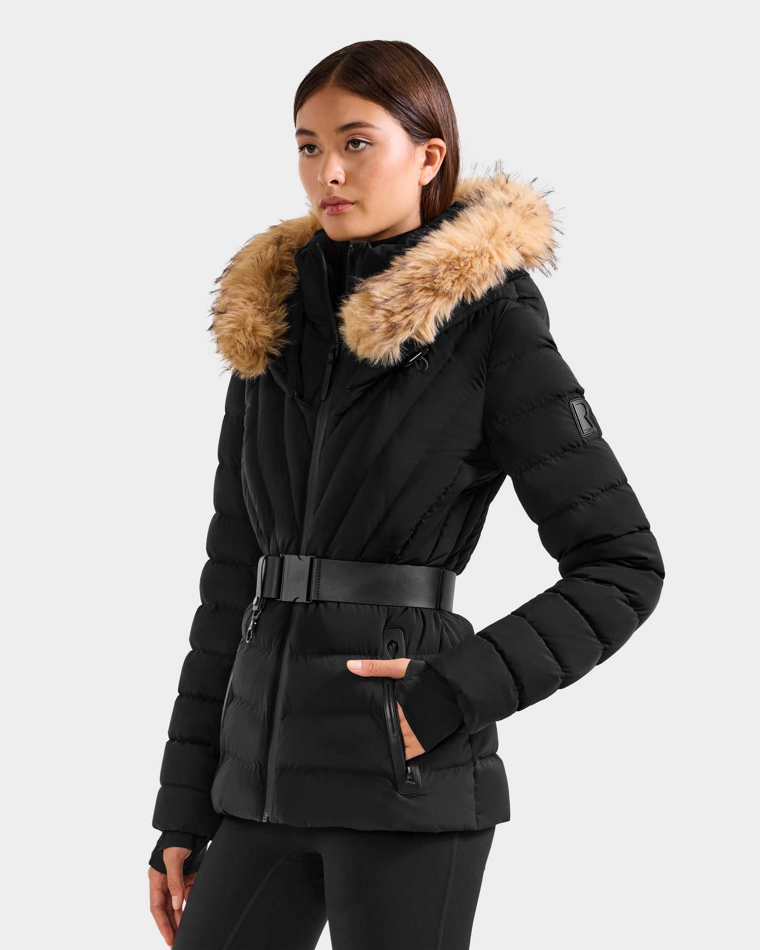 A woman wears the Rudsak ALISA F - L125527 Black belted quilted puffer jacket with RDS-certified down fill and a faux-fur trim hood, standing against a light grey background with her hands in her pockets.