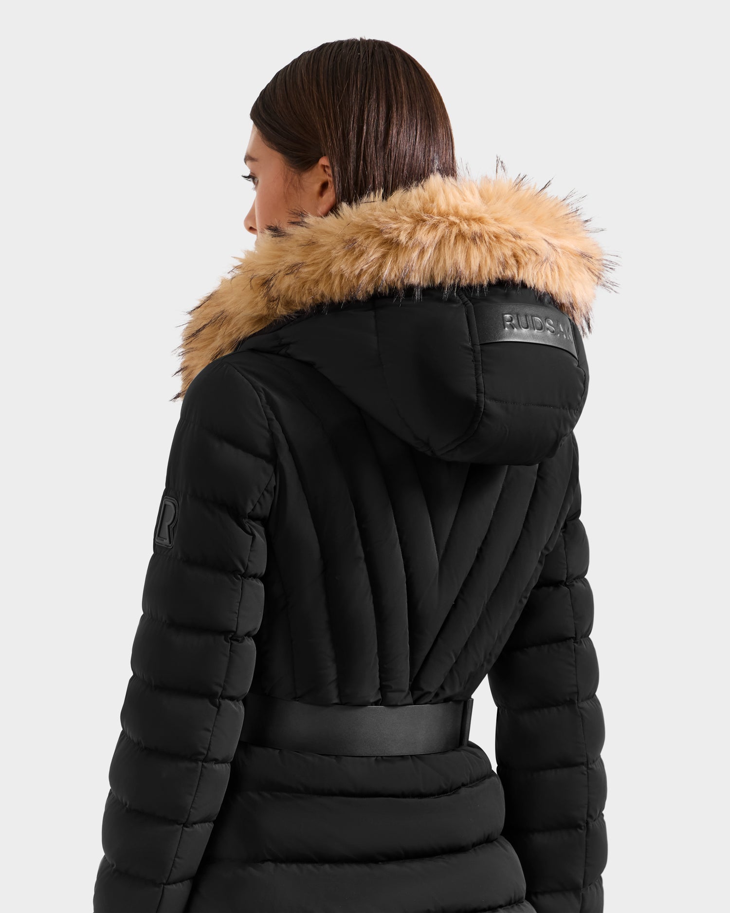 A woman with straight brown hair models the Rudsak ALISA F - L125527 Black quilted puffer coat with a belted waist and faux-fur hood, facing away from the camera against a plain light background.