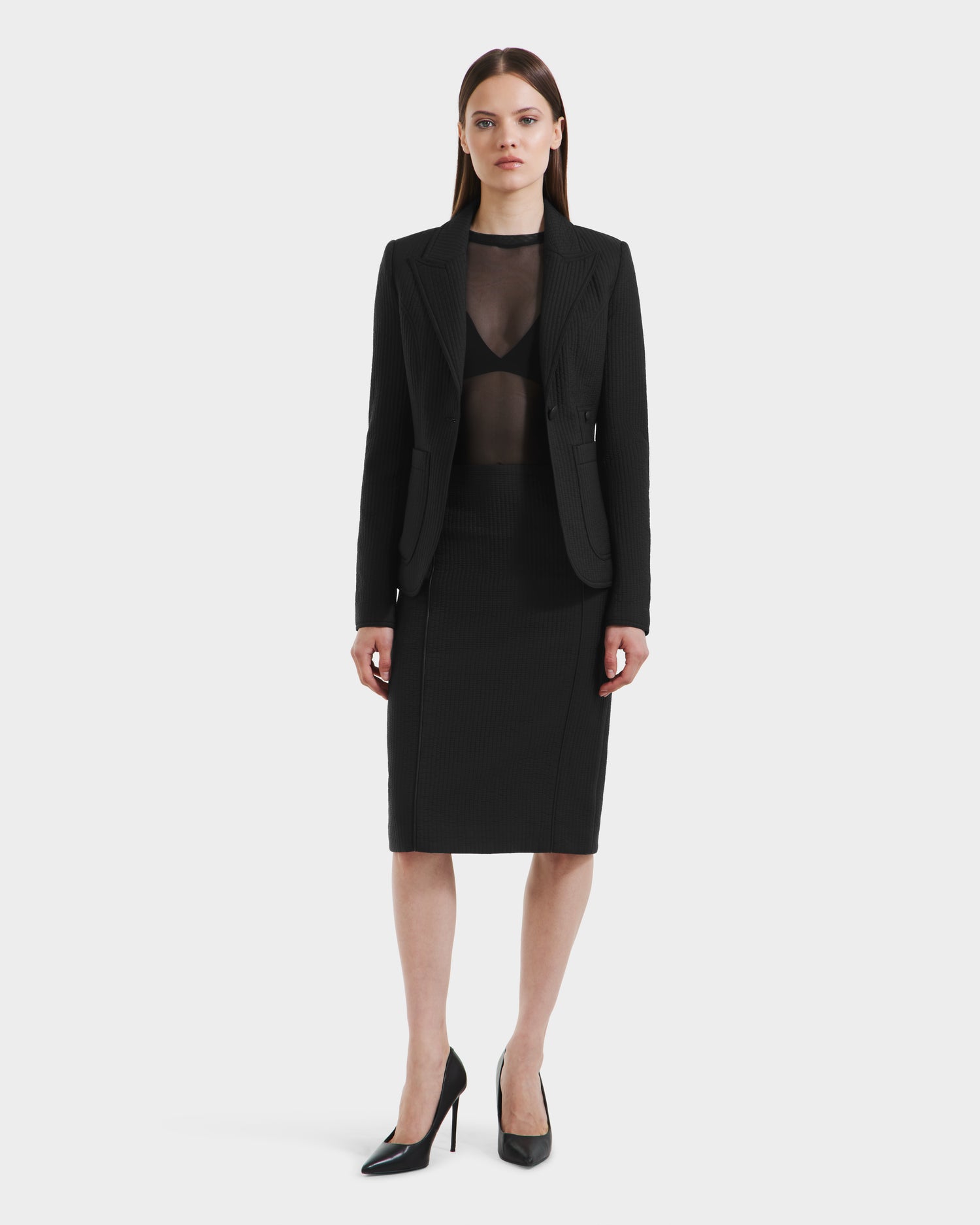 A woman faces forward against a white background, wearing the Rudsak ALVERO Q - 8525629 Black womens fitted blazer over a sheer black top, paired with a black knee-length skirt and black high-heeled shoes.