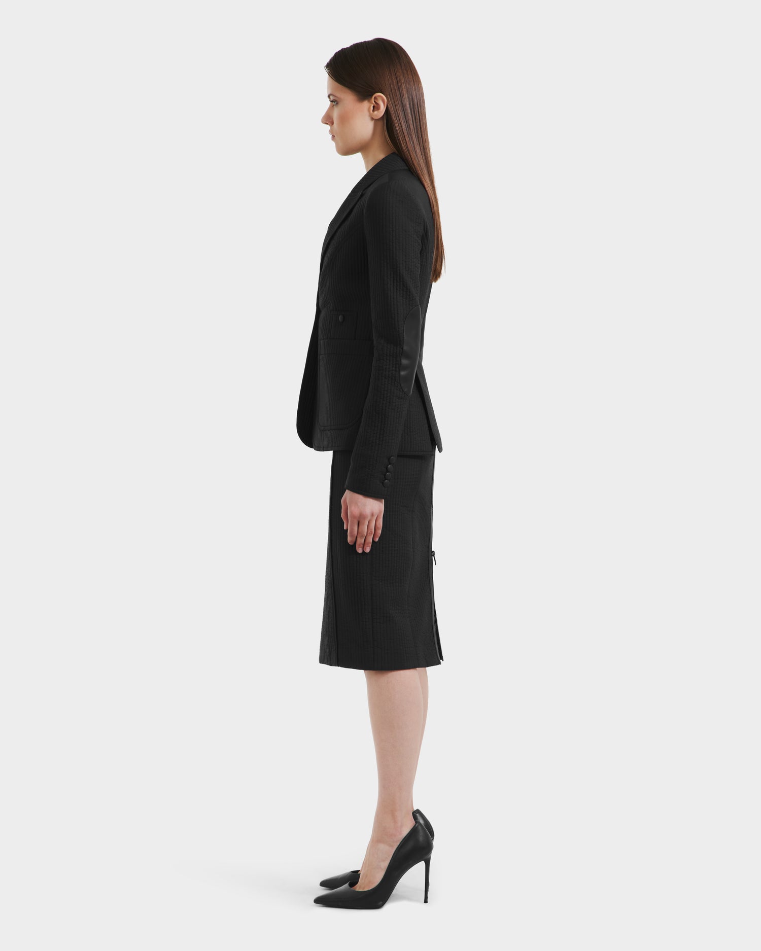 A woman with long straight brown hair, in a Rudsak ALVERO Q - 8525629 Black fitted blazer and matching knee-length skirt with black high heels, stands in profile against a plain white background.