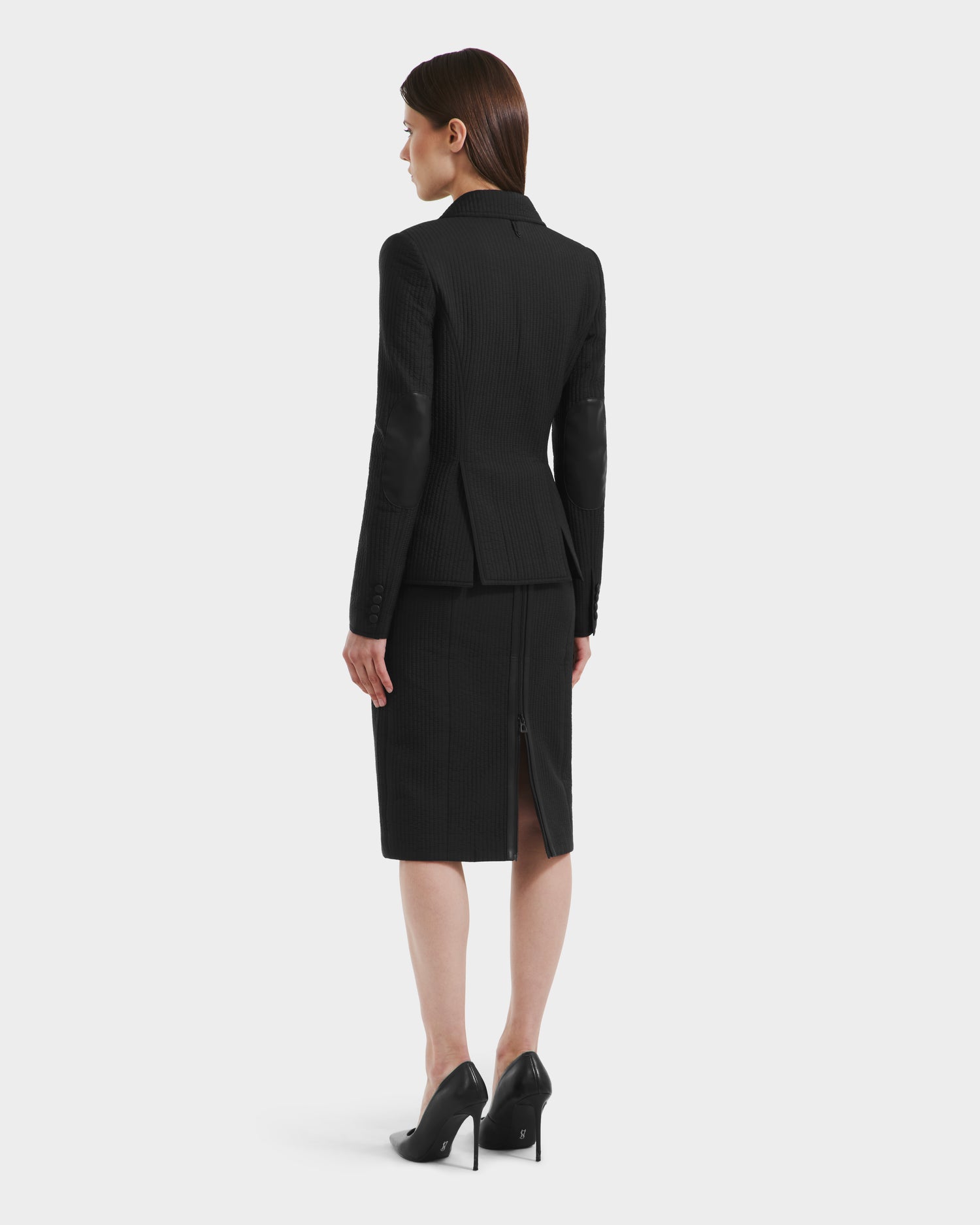 A woman with straight brown hair faces away, wearing the Rudsak ALVERO Q - 8525629 Black women’s fitted blazer and matching knee-length skirt, paired with black heels, against a plain white background.