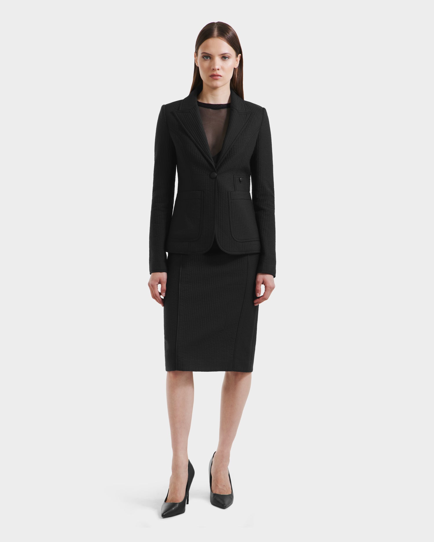 A woman stands facing forward, wearing the Rudsak ALVERO Q - 8525629 Black women’s fitted blazer over a black sheer top and matching pencil skirt, paired with black pointed-toe heels, against a plain white background.