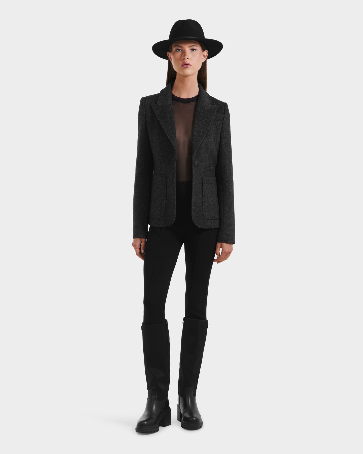 A woman stands facing forward against a white background, wearing the Rudsak ALVERO - 8525566 Grey women’s fitted blazer with vegan leather elbow patches, paired with a black wide-brimmed hat, sheer top, black pants, and knee-high boots.