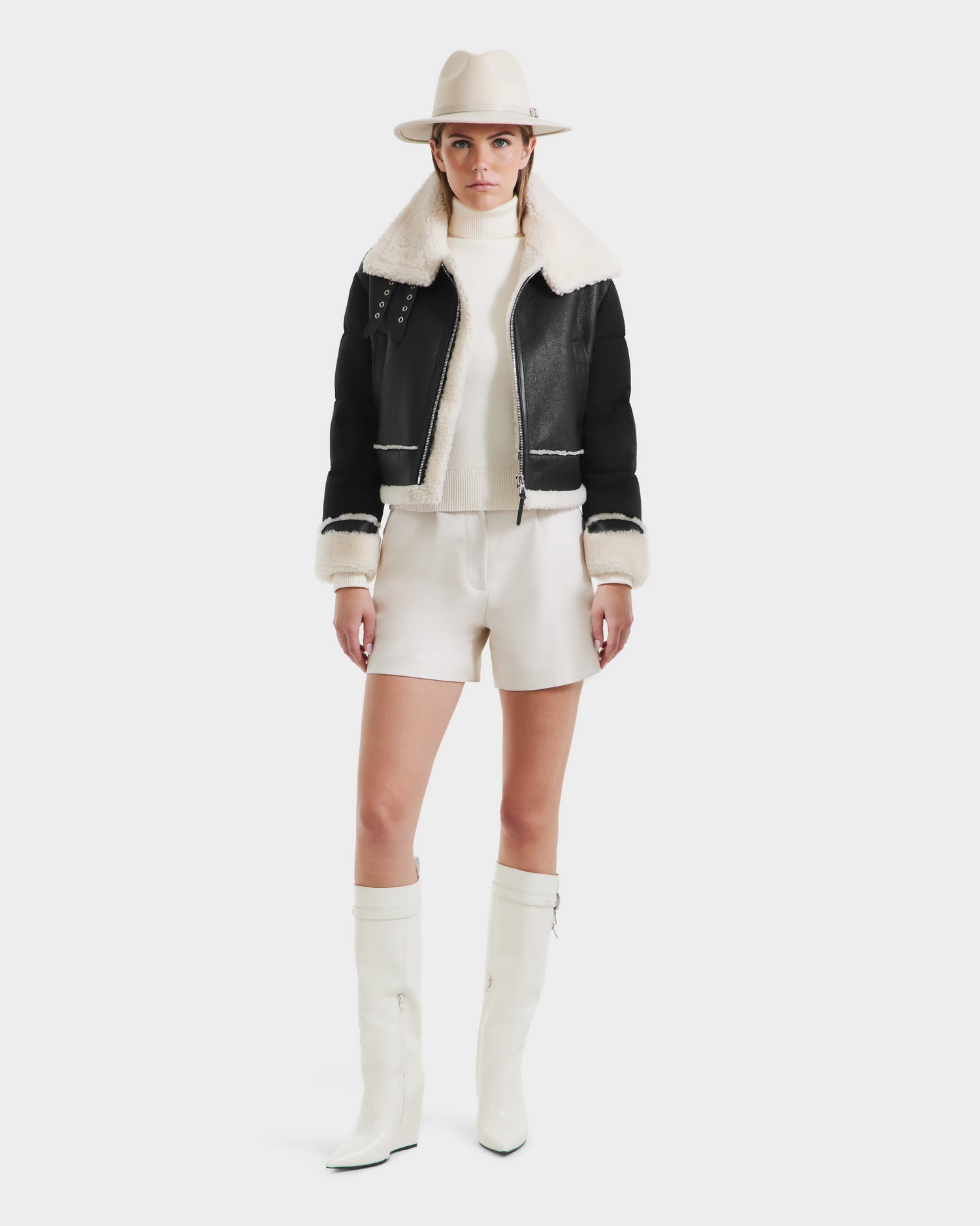 A woman wearing a beige hat, white turtleneck sweater, cream shorts, and white knee-high boots stands against a plain white background in the ANA - 8125508 Black women’s shearling jacket by Rudsak.