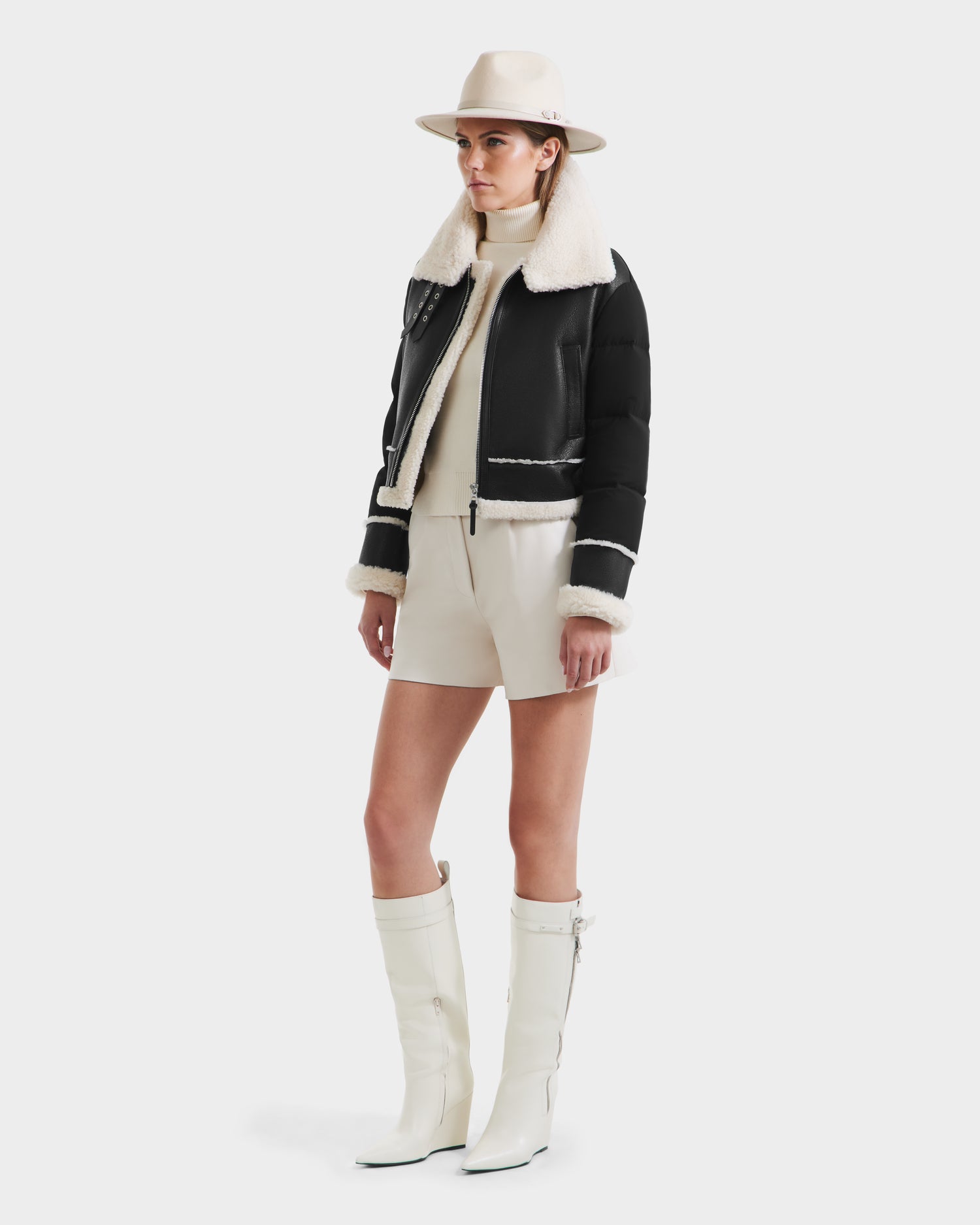 A woman stands against a white background wearing the Rudsak ANA - 8125508 Black women’s shearling jacket, a light hat, cream turtleneck sweater, cream shorts, and knee-high white boots.