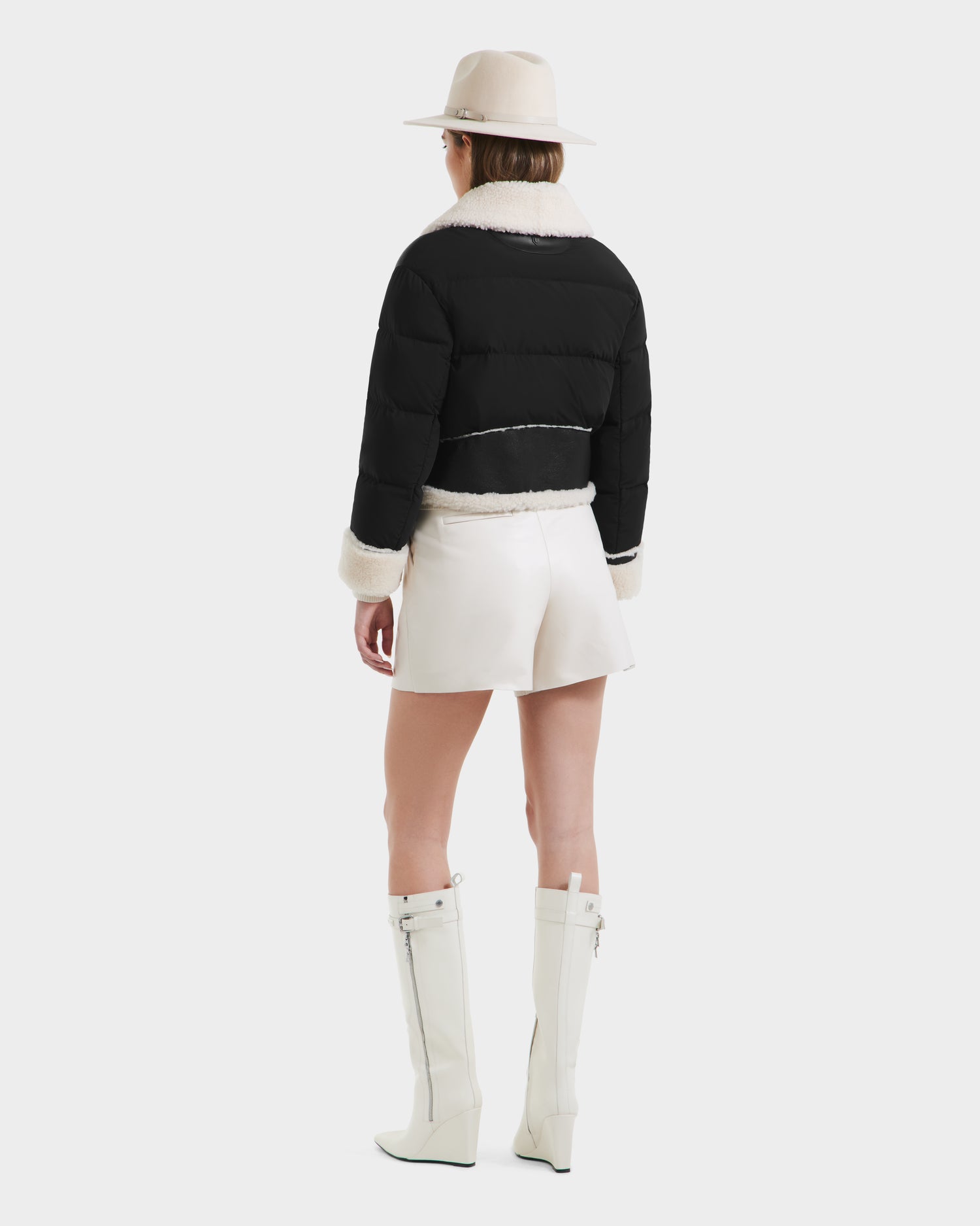 A person wearing a cream hat, white mini skirt, knee-high white boots, and the Rudsak ANA - 8125508 Black puffer jacket with white trim stands facing away against a plain background.