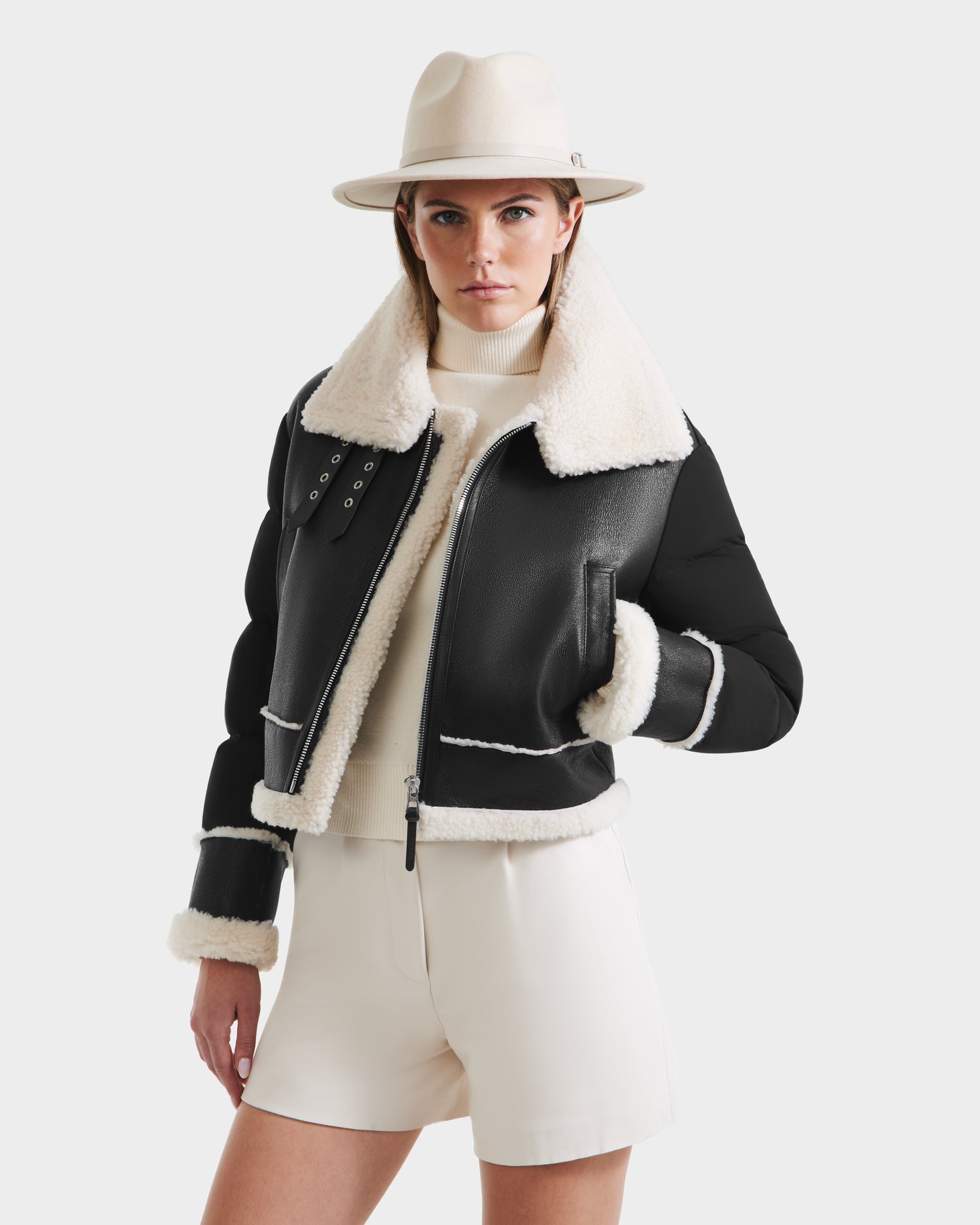 A woman wearing the Rudsak ANA - 8125508 Black womens shearling jacket with white fleece lining, a cream turtleneck sweater, and cream high-waisted shorts stands confidently against a plain white background.