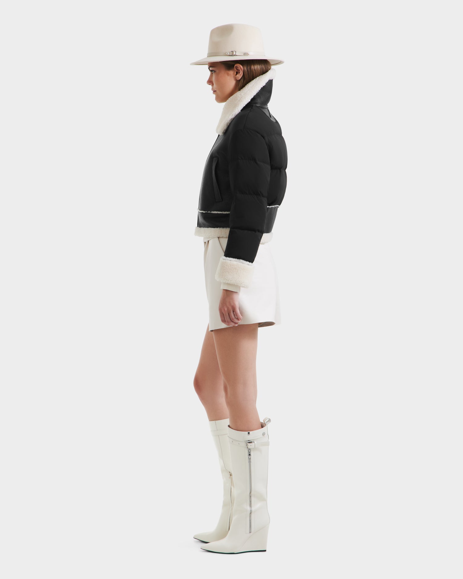 A woman in profile wears the Rudsak ANA - 8125508 Black puffer jacket with white trim, paired with white shorts, tall white heeled boots, and a cream wide-brim hat against a plain light background.