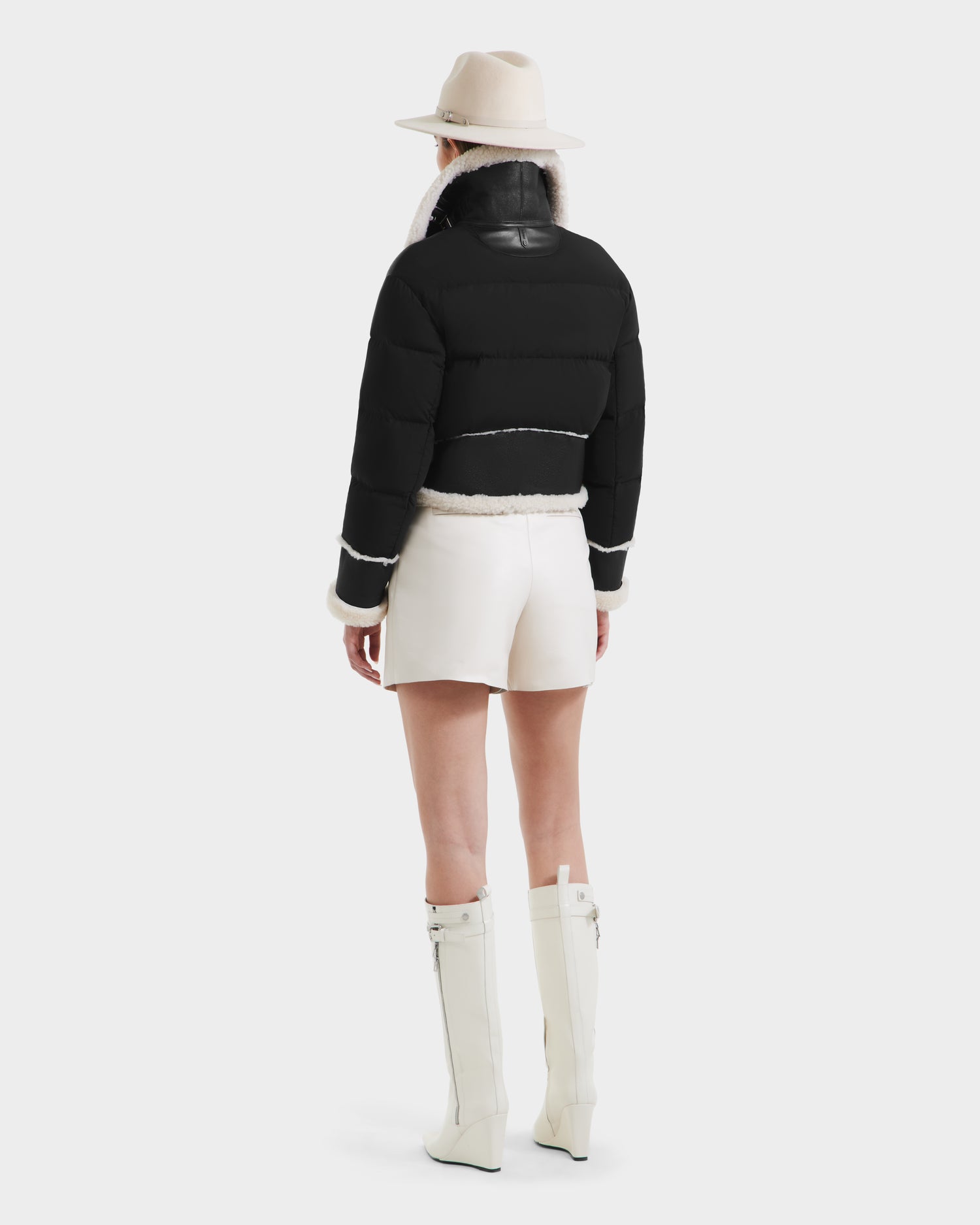 A person stands facing away against a plain background, wearing the Rudsak ANA - 8125508 Black puffer jacket with white trim, styled with white shorts, knee-high white boots, and a beige wide-brimmed hat.