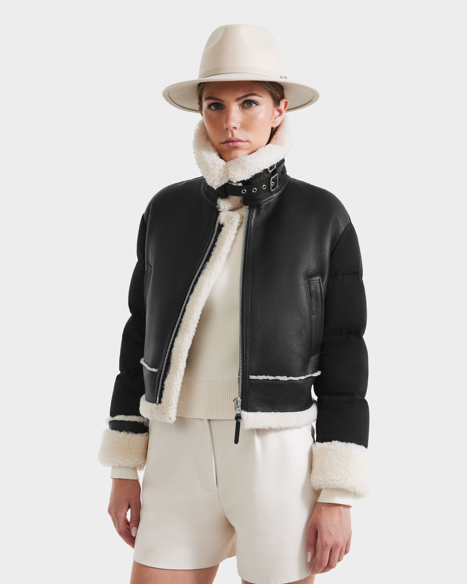 A woman wearing the Rudsak ANA - 8125508 Black women’s shearling jacket with cream lining and cuffs, a wide-brimmed beige hat, and cream shorts stands against a plain white background.
