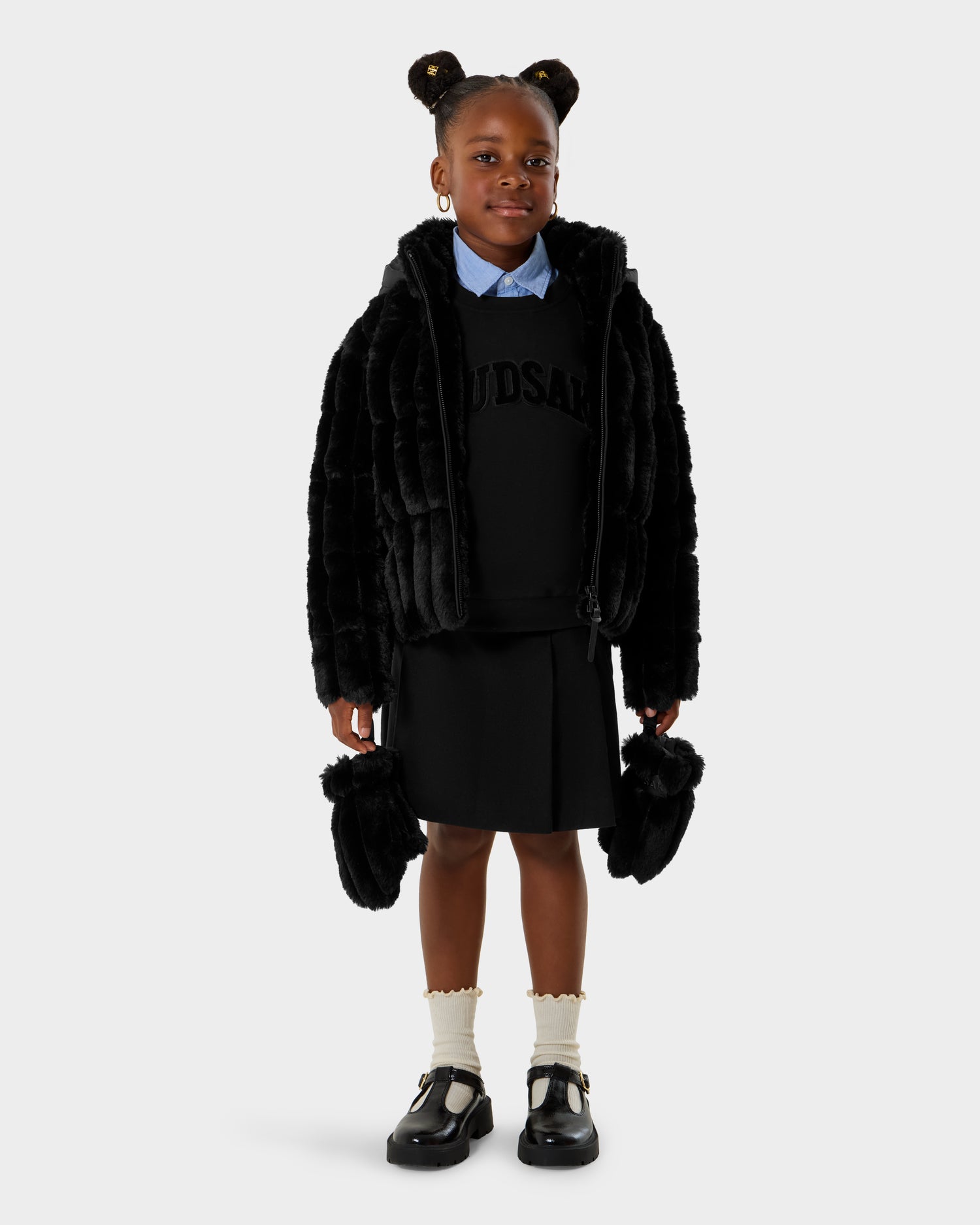 A young girl wears the Rudsak ANASTASIA K - 0125000 Black faux fur shell jacket over a black sweater and skirt, styled with a blue collared shirt, white ruffled socks, black shoes, and a matching black fur bag.